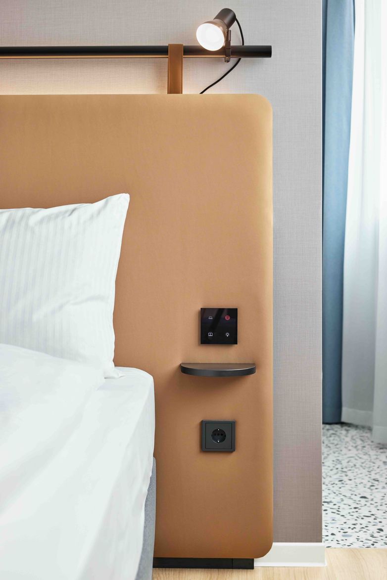 Detailaufnahme eines modernen Hotelbett-Kopfteil mit integriertem Licht, Nachtlichtschalter und Steckdose, helle Bettwäsche und Vorhänge vermitteln eine ruhige, komfortable Atmosphäre.