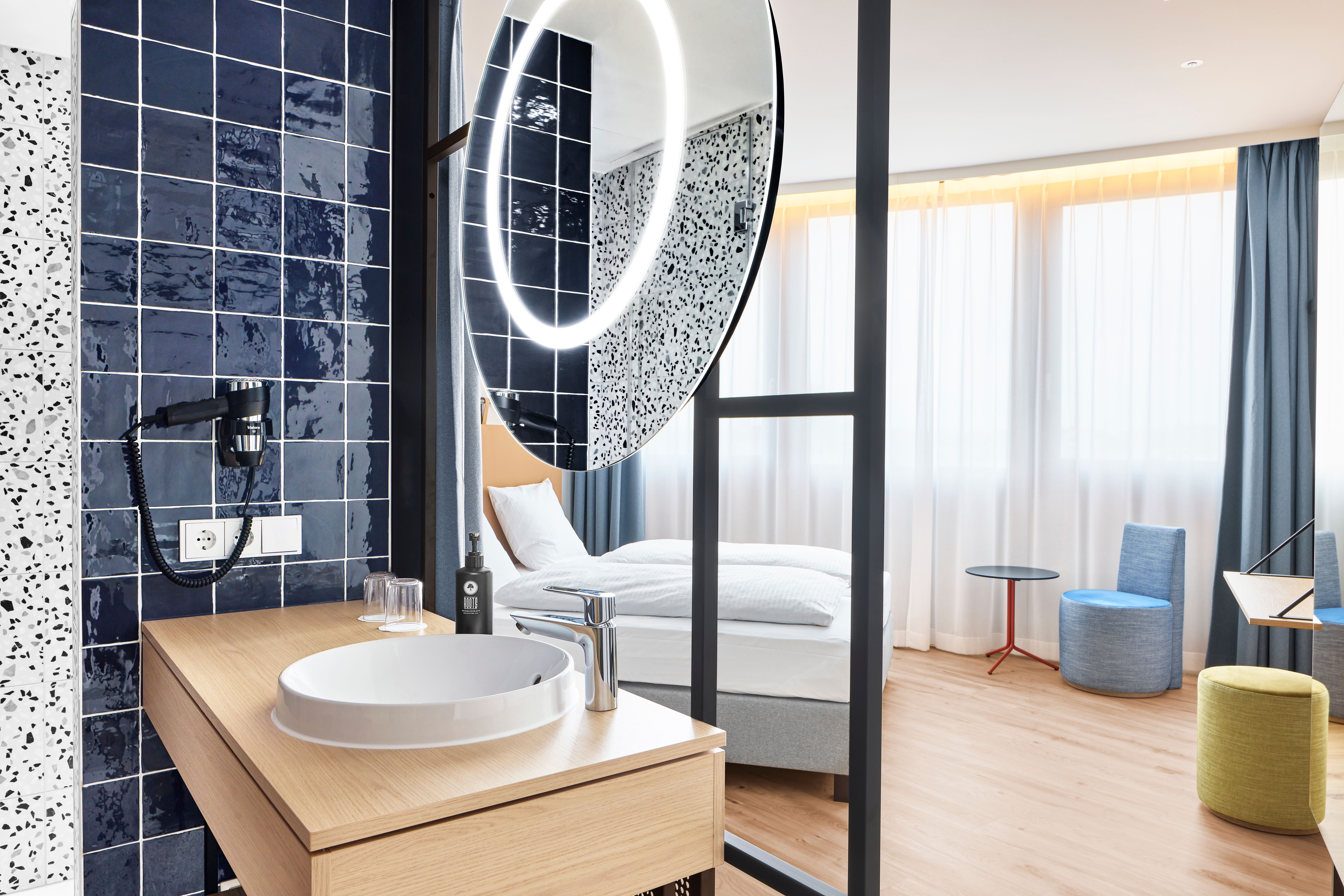 Modernes Hotelzimmer mit hellen Holzböden, gemütlichem Bett, großer Fensterfront und Vorhängen. Offenes Badezimmer mit rundem Waschbecken, beleuchtetem Spiegel und dunkelblauen Fliesen. Helle, freundliche Atmosphäre, die Komfort und Stil vereint.