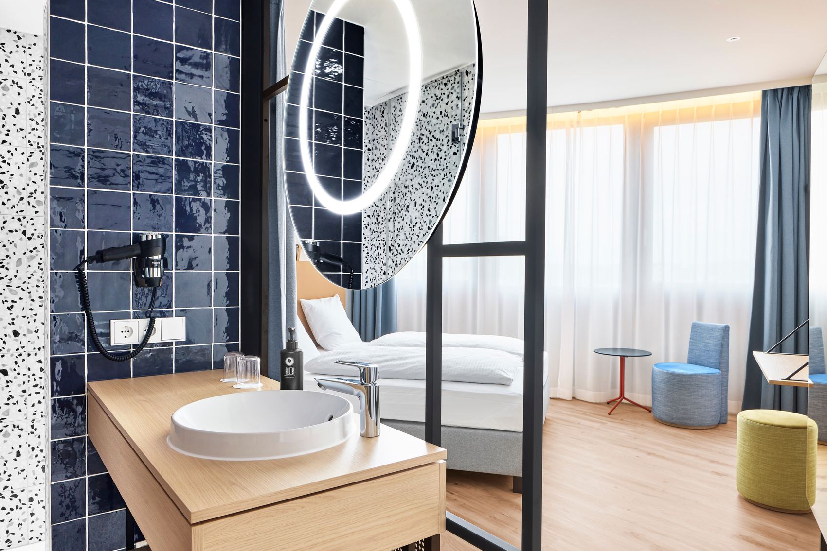Modernes Hotelzimmer mit hellen Holzböden, gemütlichem Bett, großer Fensterfront und Vorhängen. Offenes Badezimmer mit rundem Waschbecken, beleuchtetem Spiegel und dunkelblauen Fliesen. Helle, freundliche Atmosphäre, die Komfort und Stil vereint.