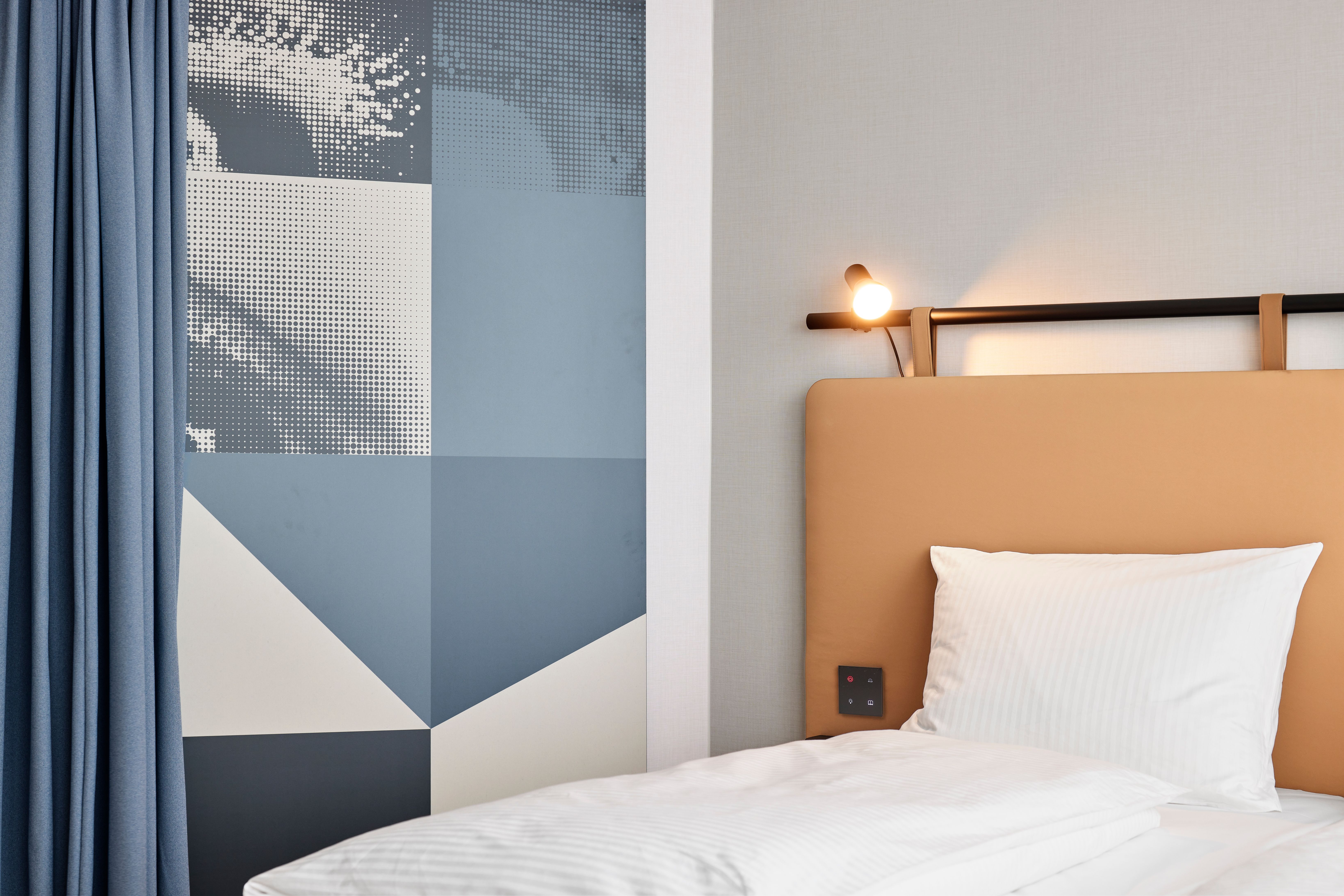 Modern eingerichtetes Hotelzimmer mit gemütlichem Einzelbett, hellen Bettwaren und einer stilvollen, geometrischen Wandgestaltung in Blau- und Grautönen neben dem Kopfteil. Ein kleiner Leselicht scheint warm über dem Bett.