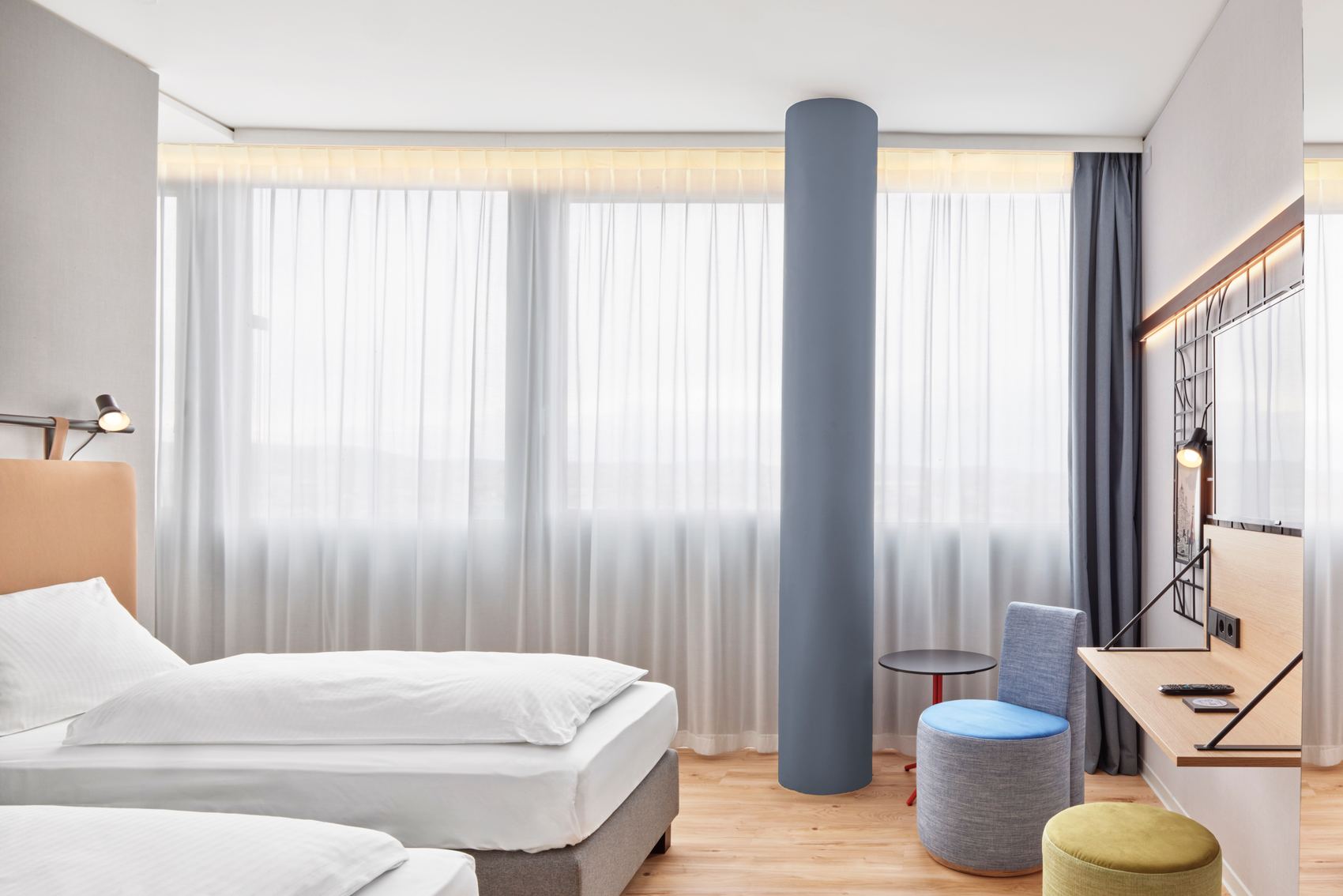 Helles Hotelzimmer mit zwei Einzelbetten, großen Fenstern mit lichtdurchlässigen Vorhängen, modernem Schreibtisch, Sitzgelegenheiten und warmem Holzboden, das eine ruhige und einladende Atmosphäre für Erholung oder Arbeit bietet.