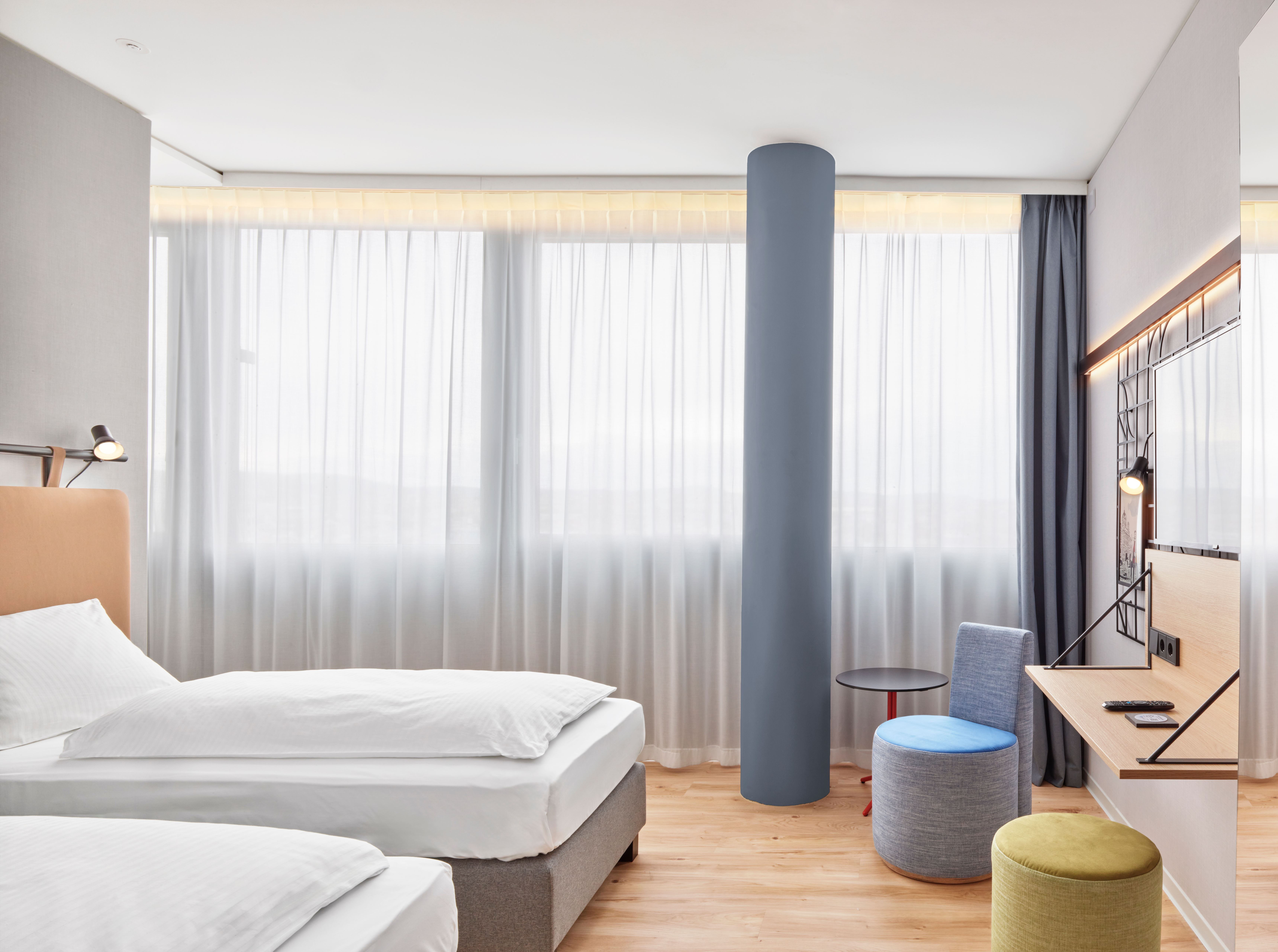 Helles Hotelzimmer mit zwei Einzelbetten, großen Fenstern mit lichtdurchlässigen Vorhängen, modernem Schreibtisch, Sitzgelegenheiten und warmem Holzboden, das eine ruhige und einladende Atmosphäre für Erholung oder Arbeit bietet. Helles Hotelzimmer mit zwei Einzelbetten, großen Fenstern mit lichtdurchlässigen Vorhängen, modernem Schreibtisch, Sitzgelegenheiten und warmem Holzboden, das eine ruhige und einladende Atmosphäre für Erholung oder Arbeit bietet.