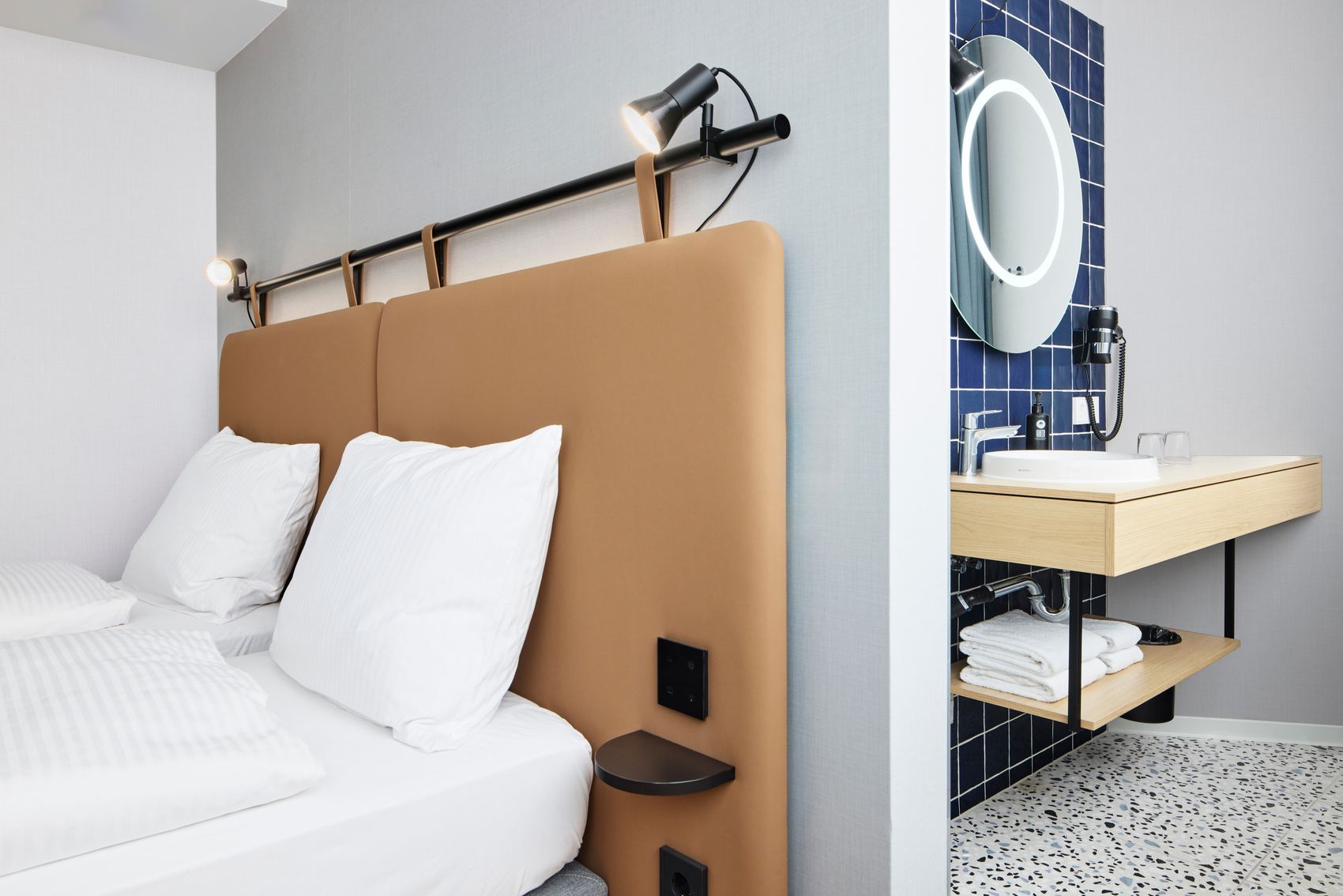 Modernes Hotelzimmer mit hellem, bequemen Doppelbett, weichen weißen Kissen und dekorativem braunem Kopfteil. Angrenzendes Badezimmer mit beleuchtetem rundem Spiegel, Holzwaschtisch und frisch gefalteten Handtüchern, das Komfort und funktionales Design vereint.
