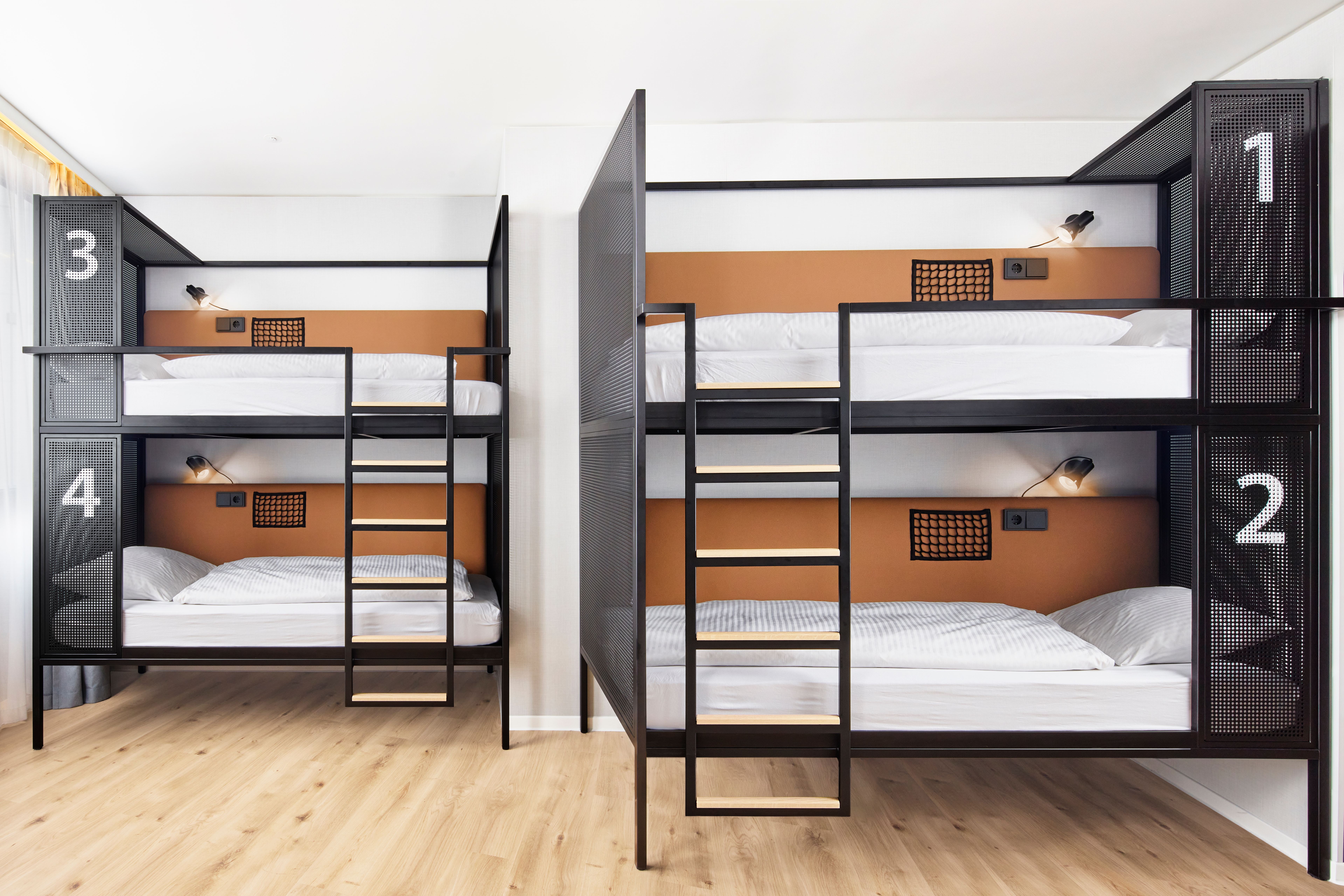 Vier moderne Etagenbetten mit klarer Nummerierung, Holzfußboden und schlichter, heller Raumgestaltung, ideal für gemeinschaftliches und funktionales Übernachten in einer Hostel- oder Jugendherbergsumgebung. Vier moderne Etagenbetten mit klarer Nummerierung, Holzfußboden und schlichter, heller Raumgestaltung, ideal für gemeinschaftliches und funktionales Übernachten in einer Hostel- oder Jugendherbergsumgebung.