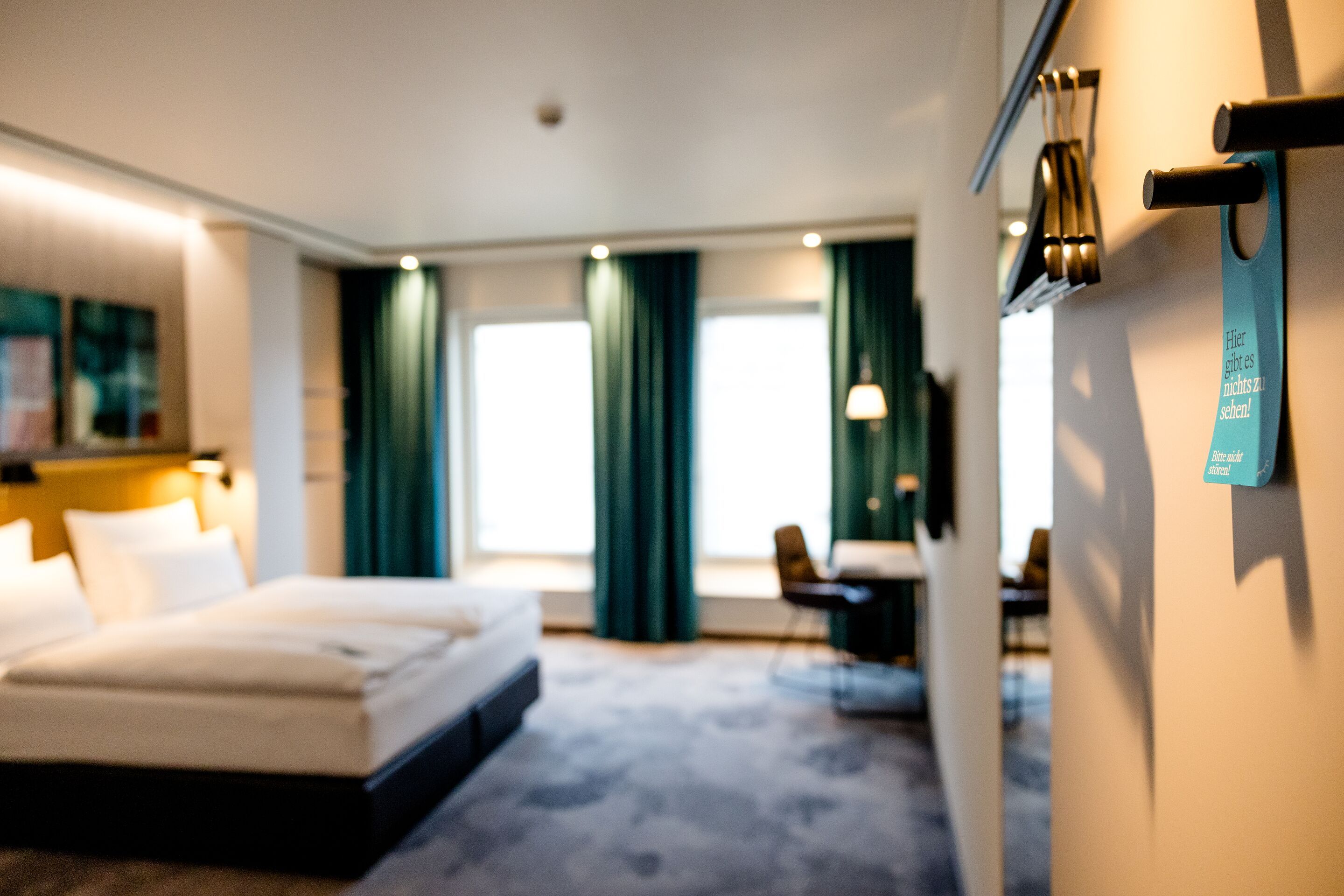 Hotel Motel One Würzburg Modern eingerichtetes Hotelzimmer mit großem Bett, angenehmer Beleuchtung und hohem Komfort. Zwei große Fenster lassen viel Tageslicht herein, Verdunkelungsvorhänge sind vorhanden. Ruhige Atmosphäre ideal für Erholung und Entspannung während des Aufenthalts.