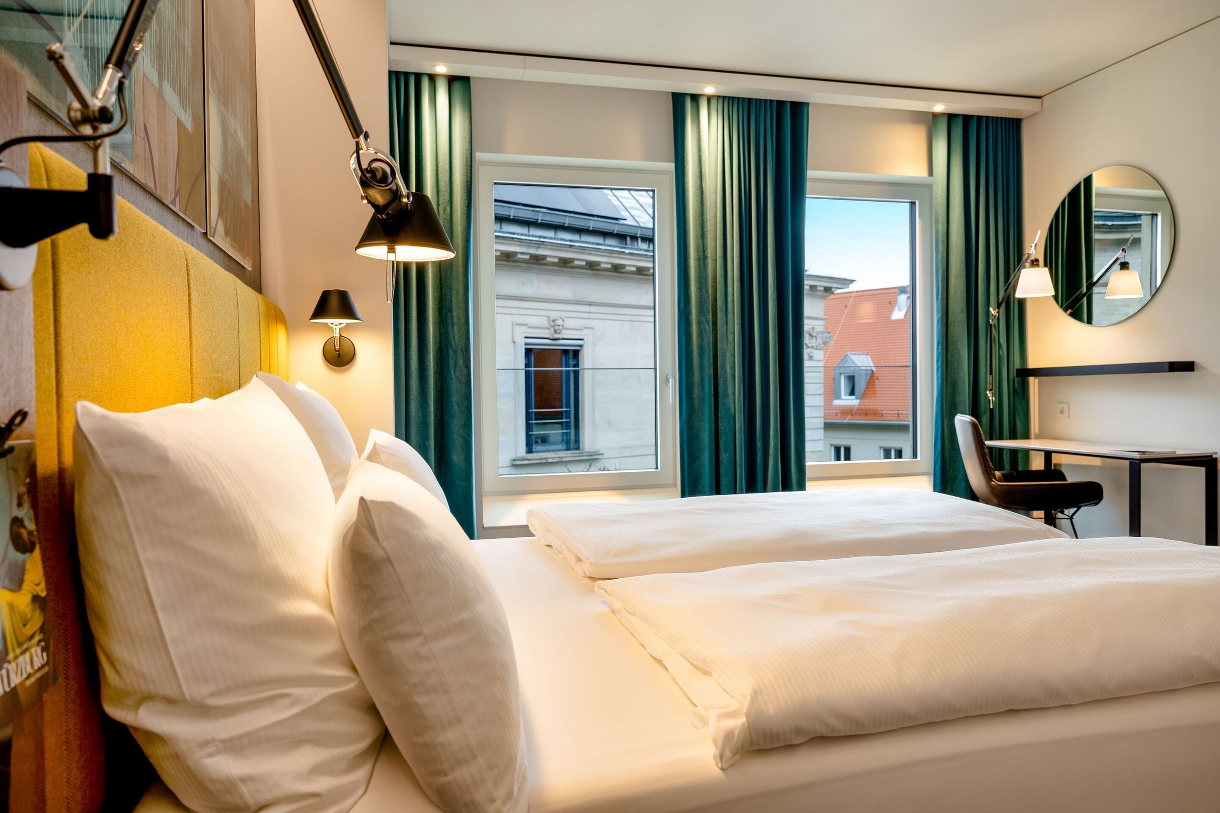 Hotel Motel One Würzburg Modernes Hotelzimmer mit Doppelbett, großen Fenstern und Blick auf historische Gebäude. Helle, freundliche Atmosphäre mit komfortabler Ausstattung, Schreibtisch und stilvollen Vorhängen. Ideal für Erholung und produktives Arbeiten.