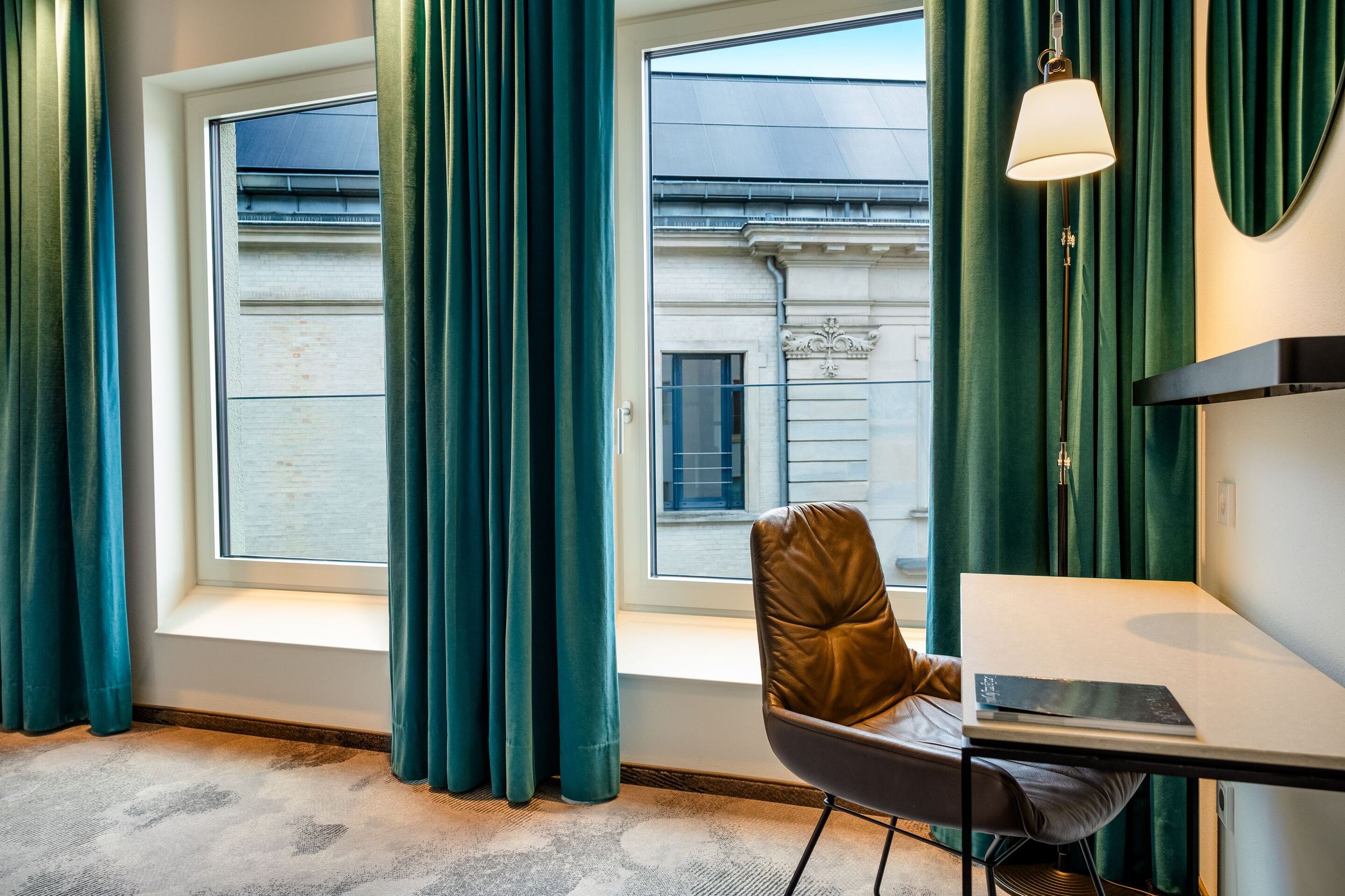 Hotel Motel One Würzburg Helles Hotelzimmer mit großen Fenstern, dunkelgrünen Vorhängen, einem Schreibtisch mit Stuhl und einer Wandlampe, das eine ruhige Arbeitsatmosphäre bietet. Blick auf ein klassisches Gebäude von außen.