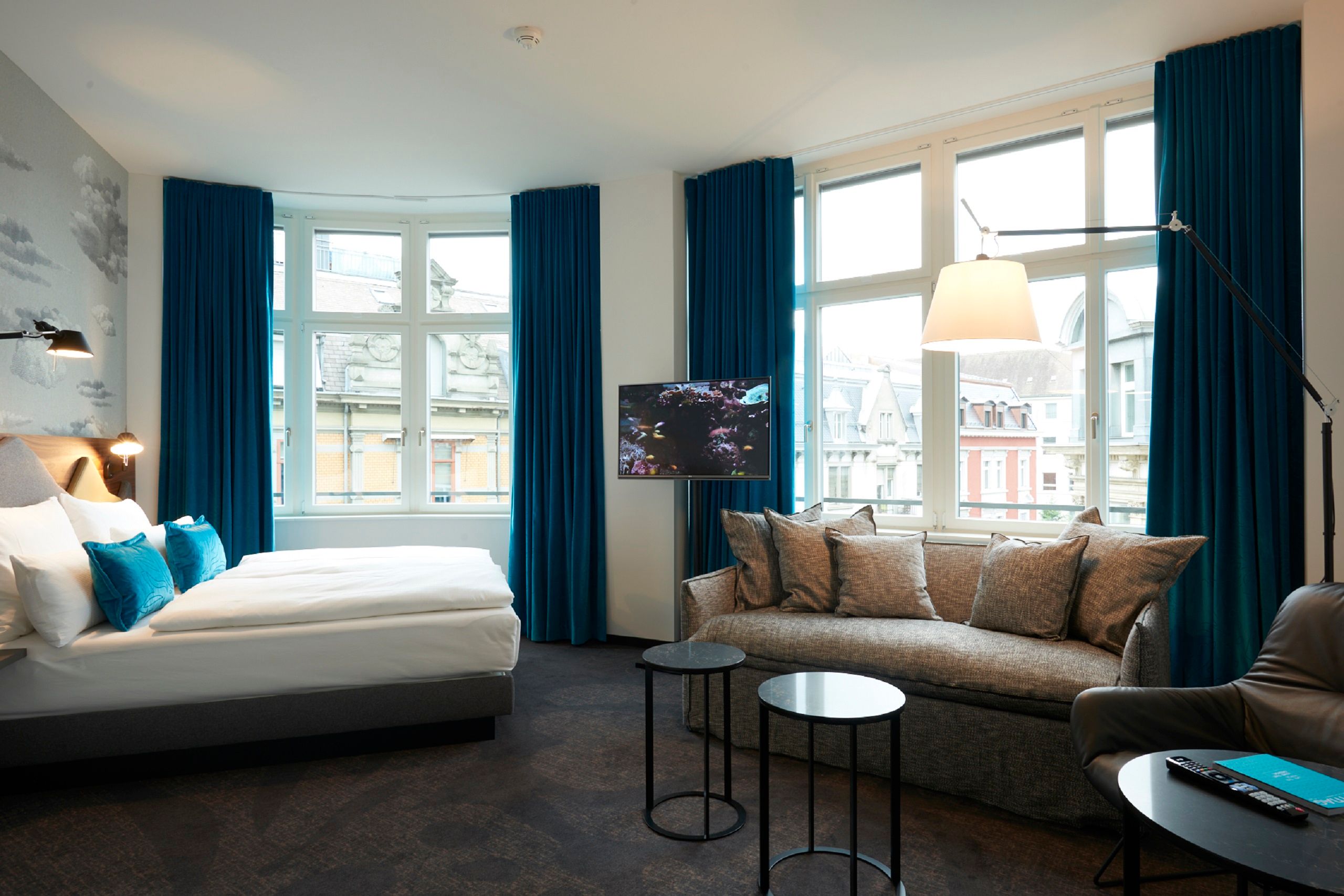 Helles Hotelzimmer mit großem Bett, gemütlichem Sofa, zwei kleinen Tischen und breiten Fenstern mit Blick auf die Stadt, dekoriert mit blauen Vorhängen und stilvoller Beleuchtung für entspannte Atmosphäre.