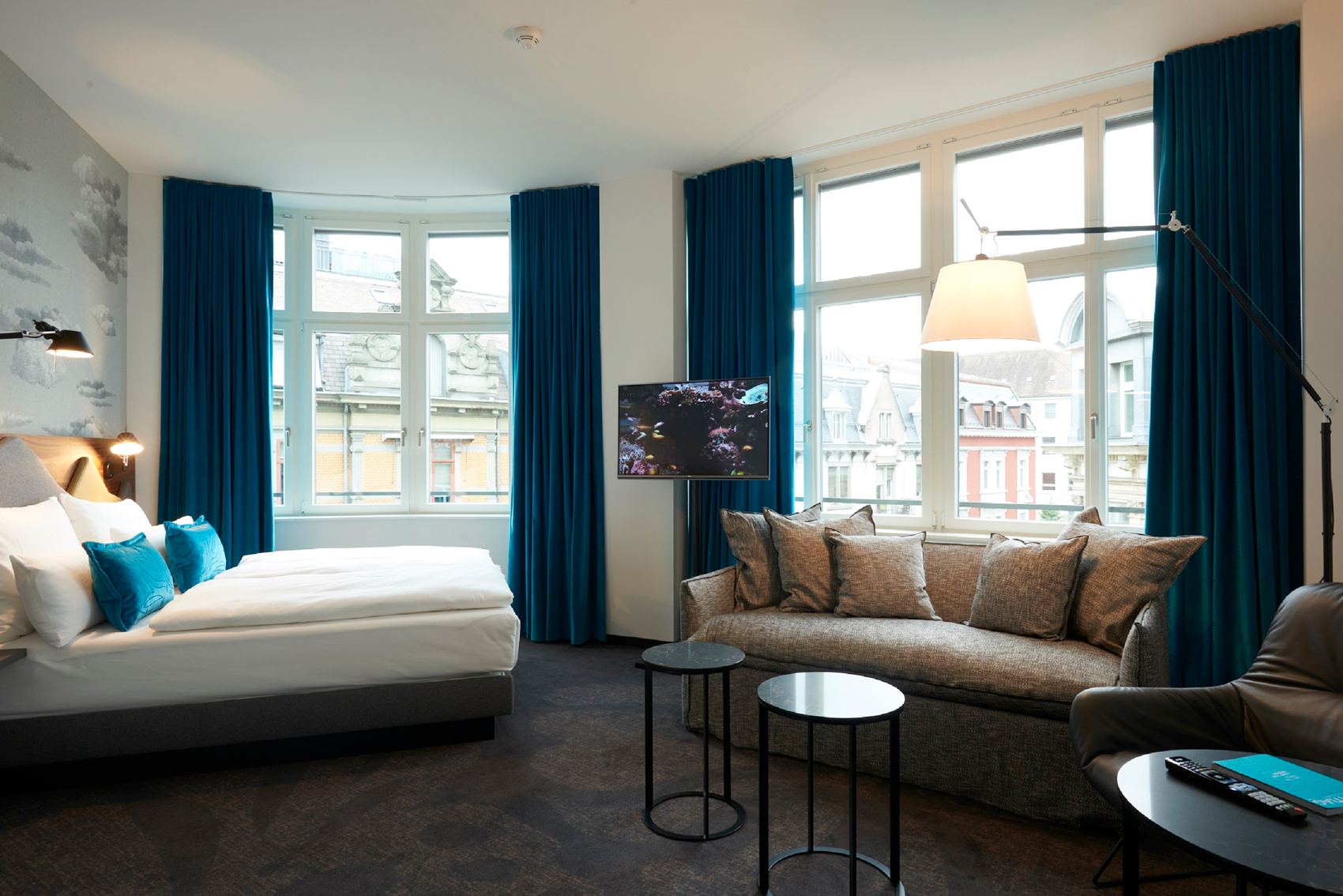Helles Hotelzimmer mit großem Bett, gemütlichem Sofa, zwei kleinen Tischen und breiten Fenstern mit Blick auf die Stadt, dekoriert mit blauen Vorhängen und stilvoller Beleuchtung für entspannte Atmosphäre.