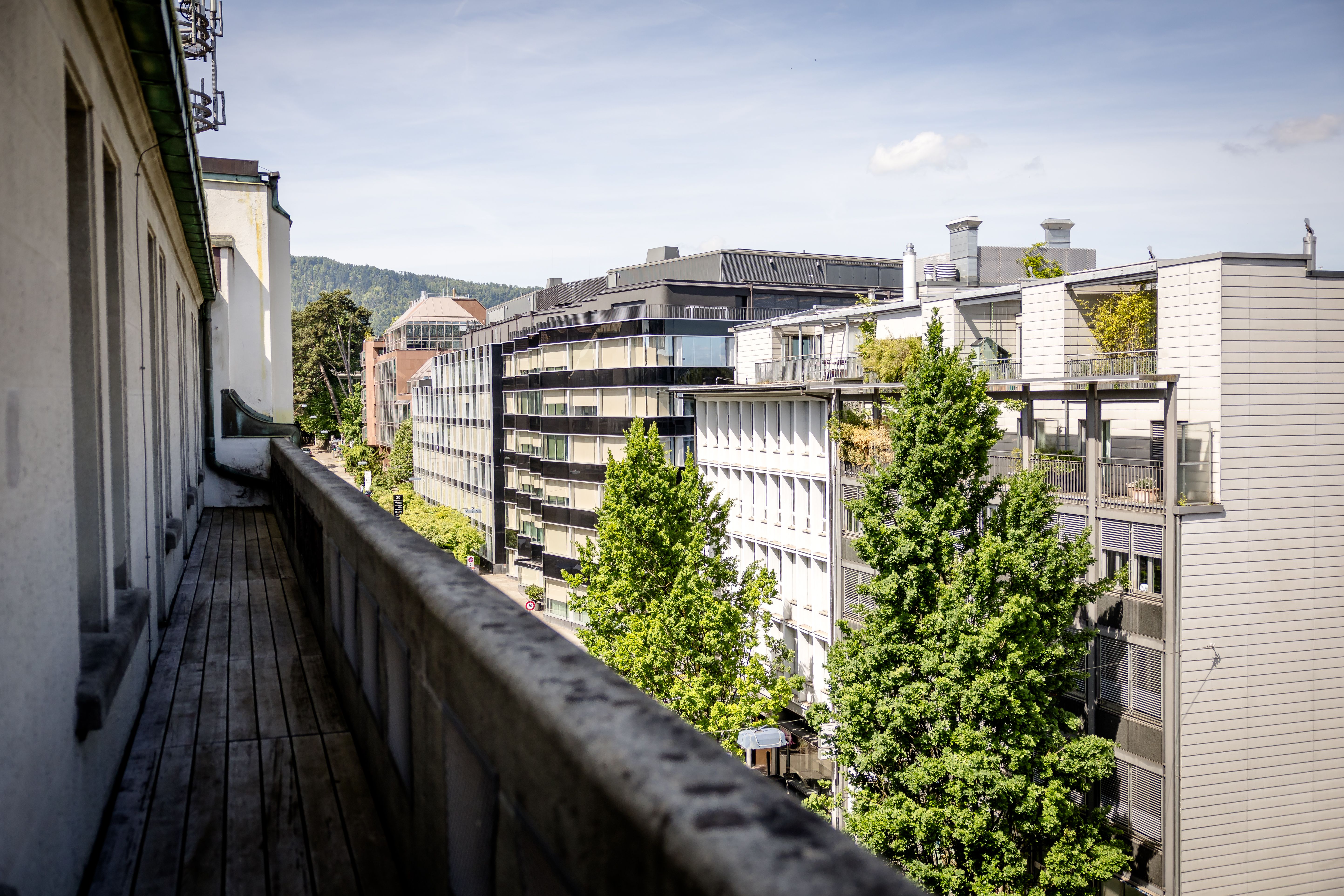 Blick von einem langen Balkon auf moderne Wohn- und Bürogebäude, umgeben von grünen Bäumen und sanften Hügeln unter blauem Himmel.