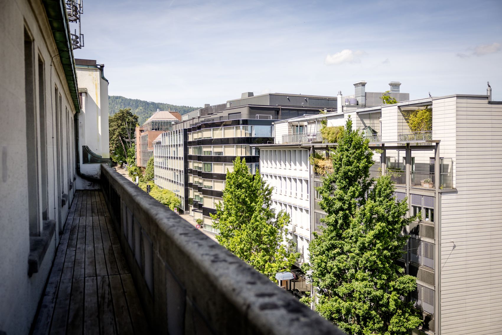 Blick von einem langen Balkon auf moderne Wohn- und Bürogebäude, umgeben von grünen Bäumen und sanften Hügeln unter blauem Himmel.