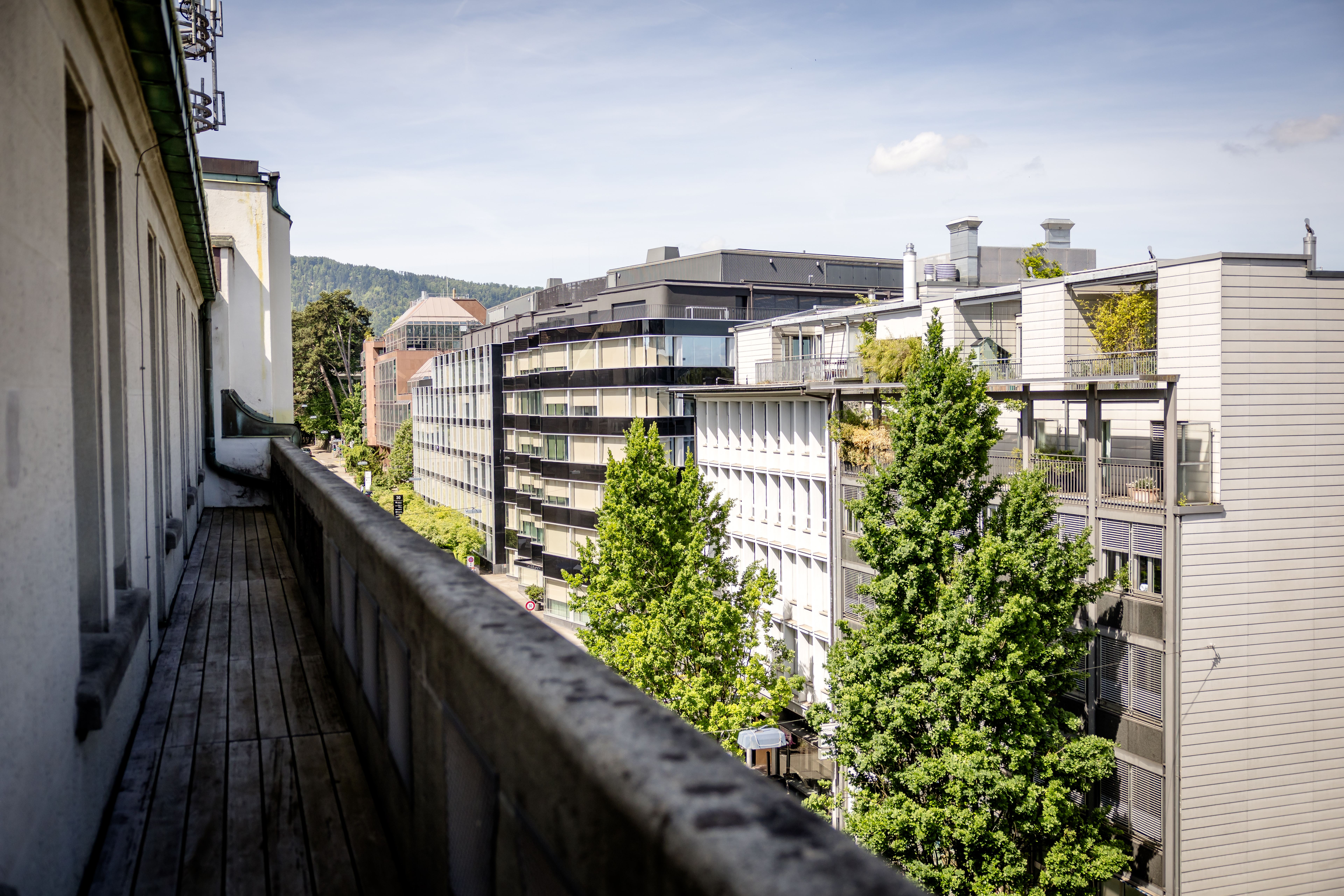 Blick von einem langen Balkon auf moderne Wohn- und Bürogebäude, umgeben von grünen Bäumen und sanften Hügeln unter blauem Himmel. Blick von einem langen Balkon auf moderne Wohn- und Bürogebäude, umgeben von grünen Bäumen und sanften Hügeln unter blauem Himmel.