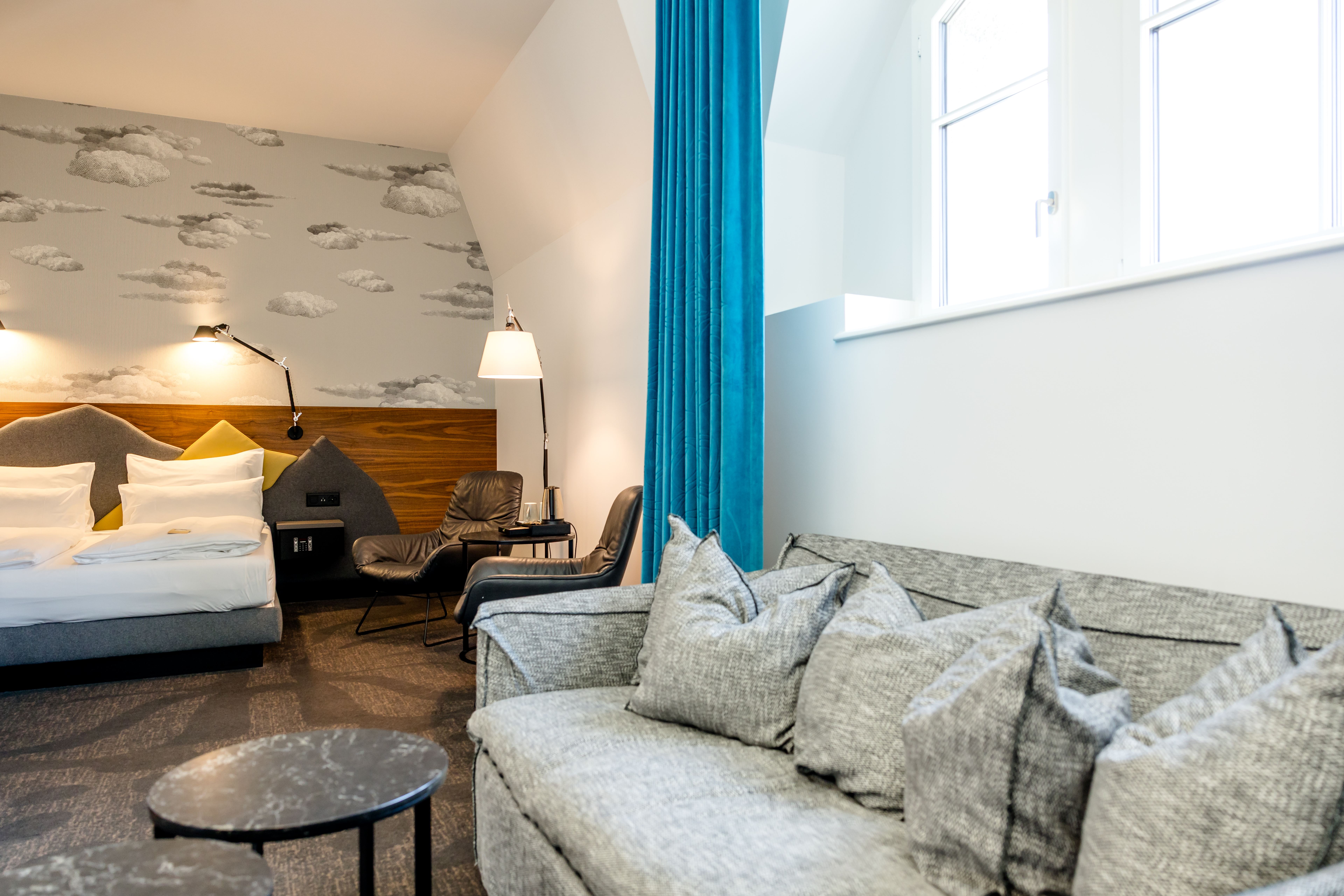 Helles Hotelzimmer mit gemütlichem Doppelbett, Sitzbereich mit grauem Sofa und Sesseln, moderner Wandgestaltung mit Wolkenmotiv und großen Fenstern, die viel Tageslicht hereinlassen. Helles Hotelzimmer mit gemütlichem Doppelbett, Sitzbereich mit grauem Sofa und Sesseln, moderner Wandgestaltung mit Wolkenmotiv und großen Fenstern, die viel Tageslicht hereinlassen.