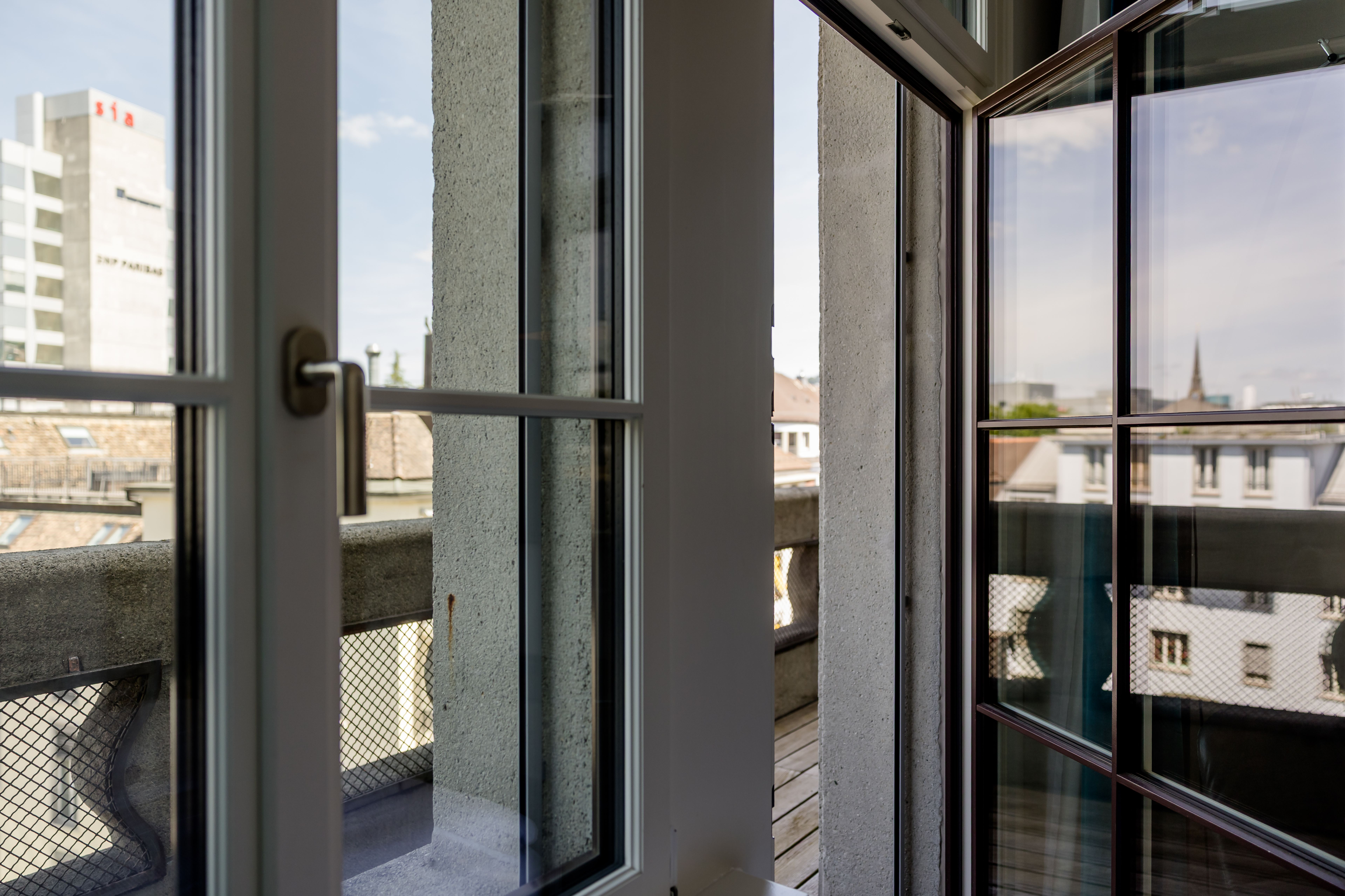 Blick von einem Zimmer auf einen Balkon mit Holzboden und Metallschutz, dahinter Stadtgebäude und Himmel bei Tageslicht. Große Fenster lassen viel natürliches Licht herein. Blick von einem Zimmer auf einen Balkon mit Holzboden und Metallschutz, dahinter Stadtgebäude und Himmel bei Tageslicht. Große Fenster lassen viel natürliches Licht herein.