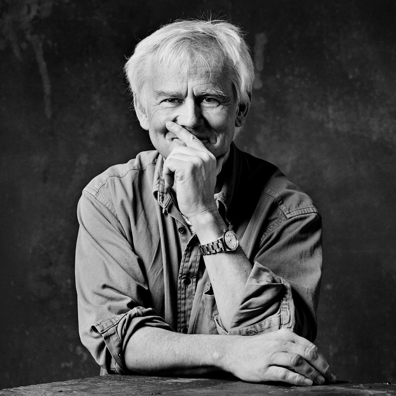 Älterer Mann mit weißem Haar sitzt entspannt, stützt das Kinn auf die Hand und blickt nachdenklich in die Kamera vor dunklem Hintergrund.