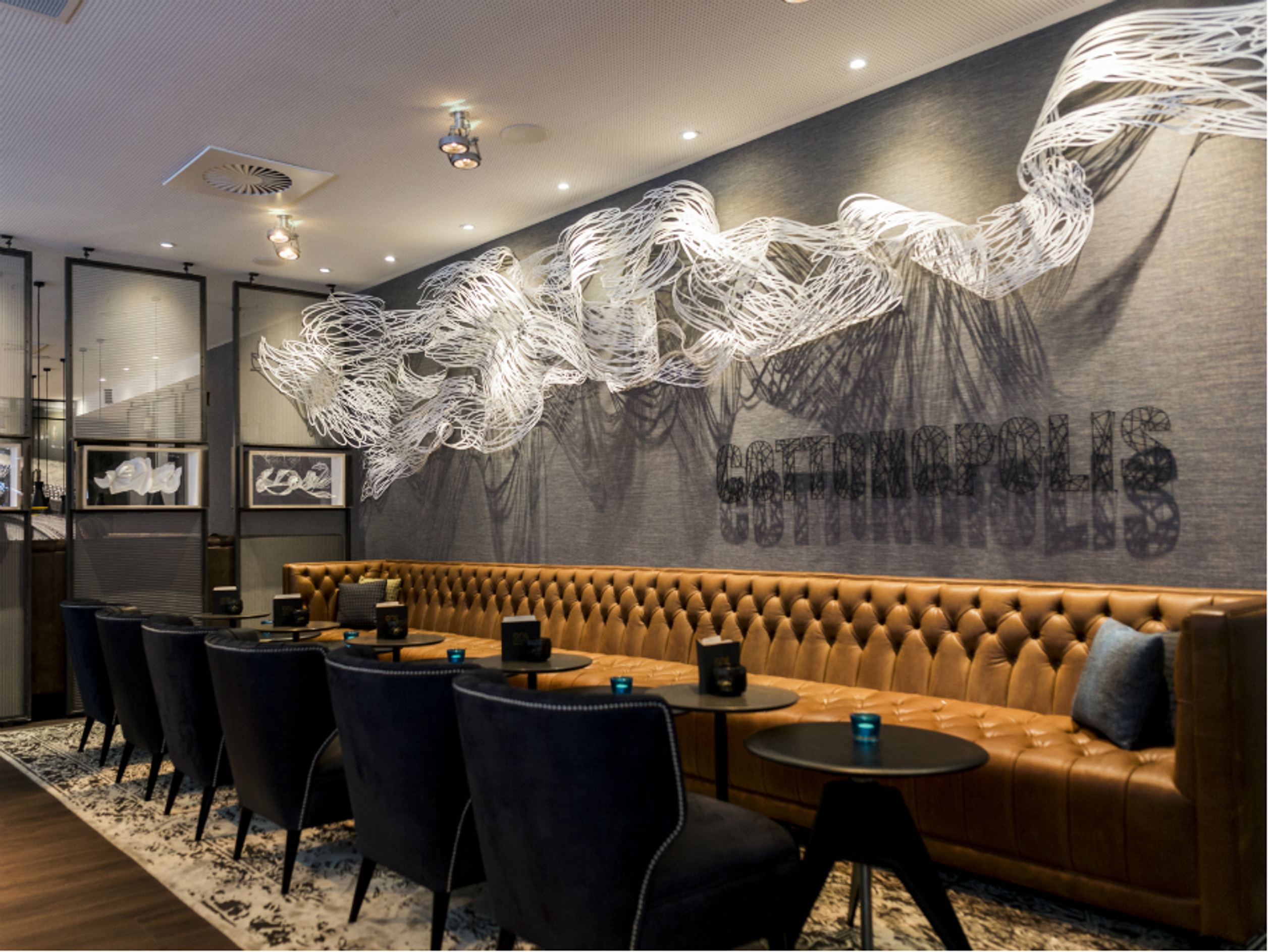 Motel One Hotel-Loungebereich mit braunem Ledersofa, schwarzen Sesseln, runden Tischen und auffälliger kunstvoller Wanddeko in Wellenform.
