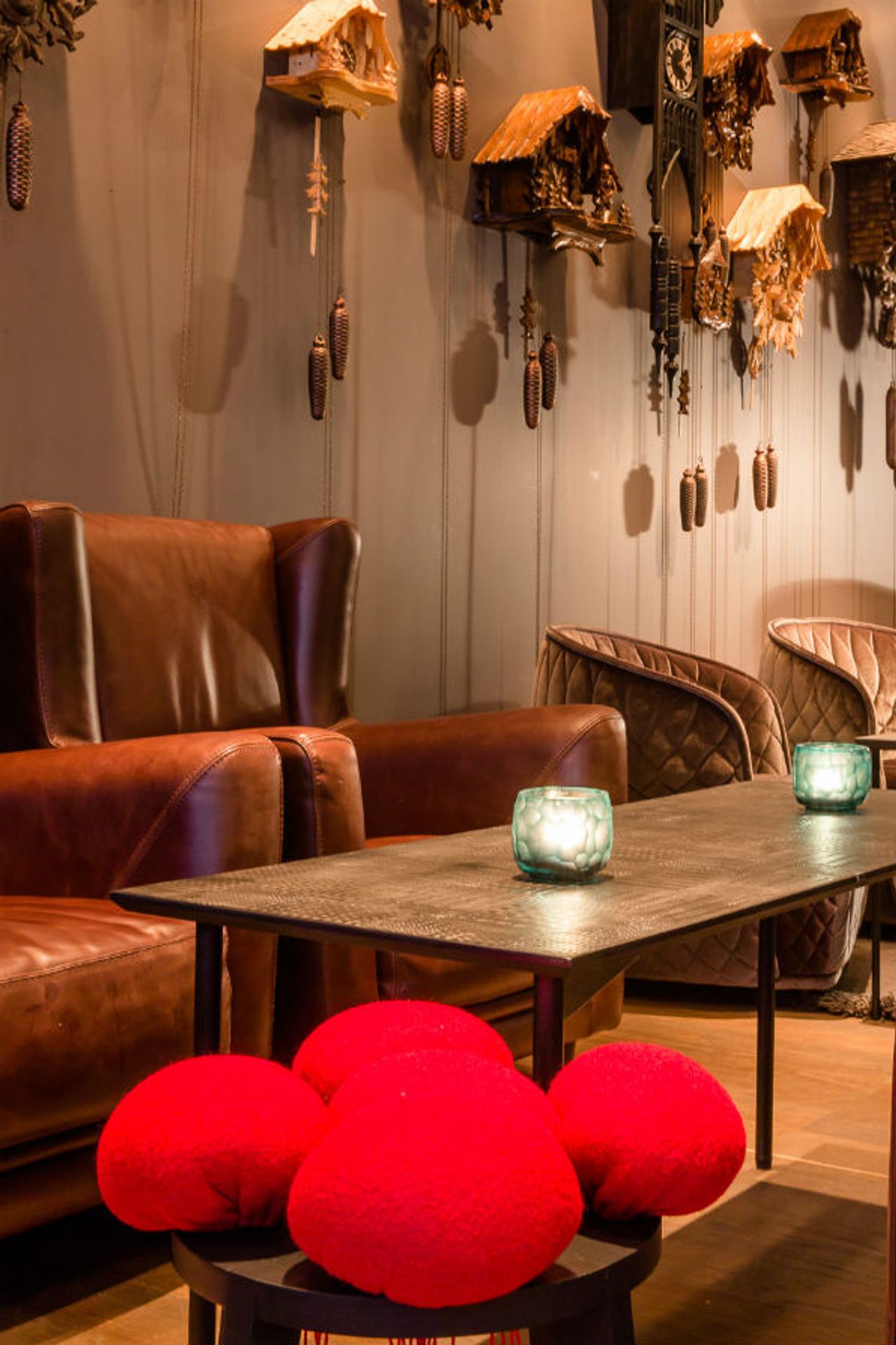 Gemütliche Motel One Lounge mit braunen Ledersesseln, einem Holztisch und dekorativen roten Kissen. An der Wand hängen kunstvolle Kuckucksuhren, die eine warme und traditionelle Atmosphäre schaffen. Zwei Kerzen auf dem Tisch sorgen für stimmungsvolles Licht.