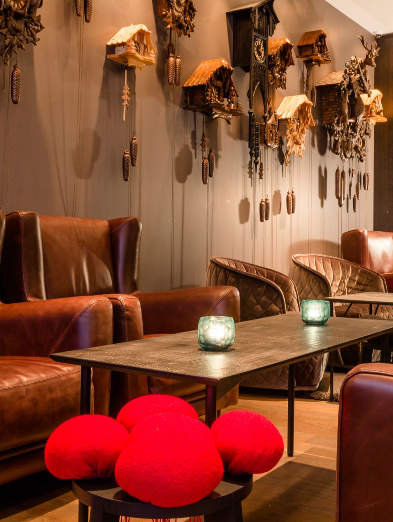 Gemütliche Motel One Sitzecke mit braunen Ledersesseln und einem Holztisch, dekoriert mit leuchtend roten Kissen. An der Wand zahlreiche kunstvolle Kuckucksuhren.