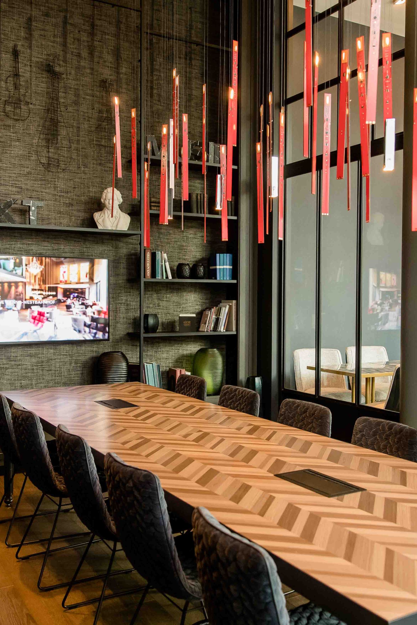 Motel One Besprechungsraum mit langem Holztisch, bequemen Stühlen und hängender LED-Beleuchtung. Große Fenster sorgen für viel Tageslicht, Bücherregale und ein Bildschirm an der Wand.