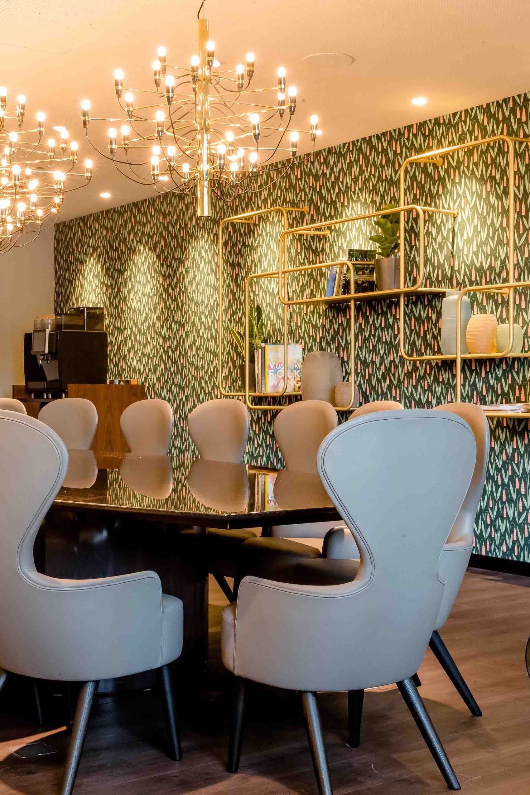 Motel One Konferenzraum mit großem dunklem Tisch, bequemen grauen Polstersesseln, dekorativer Wandgestaltung und warmem Licht durch stilvolle Kronleuchter.