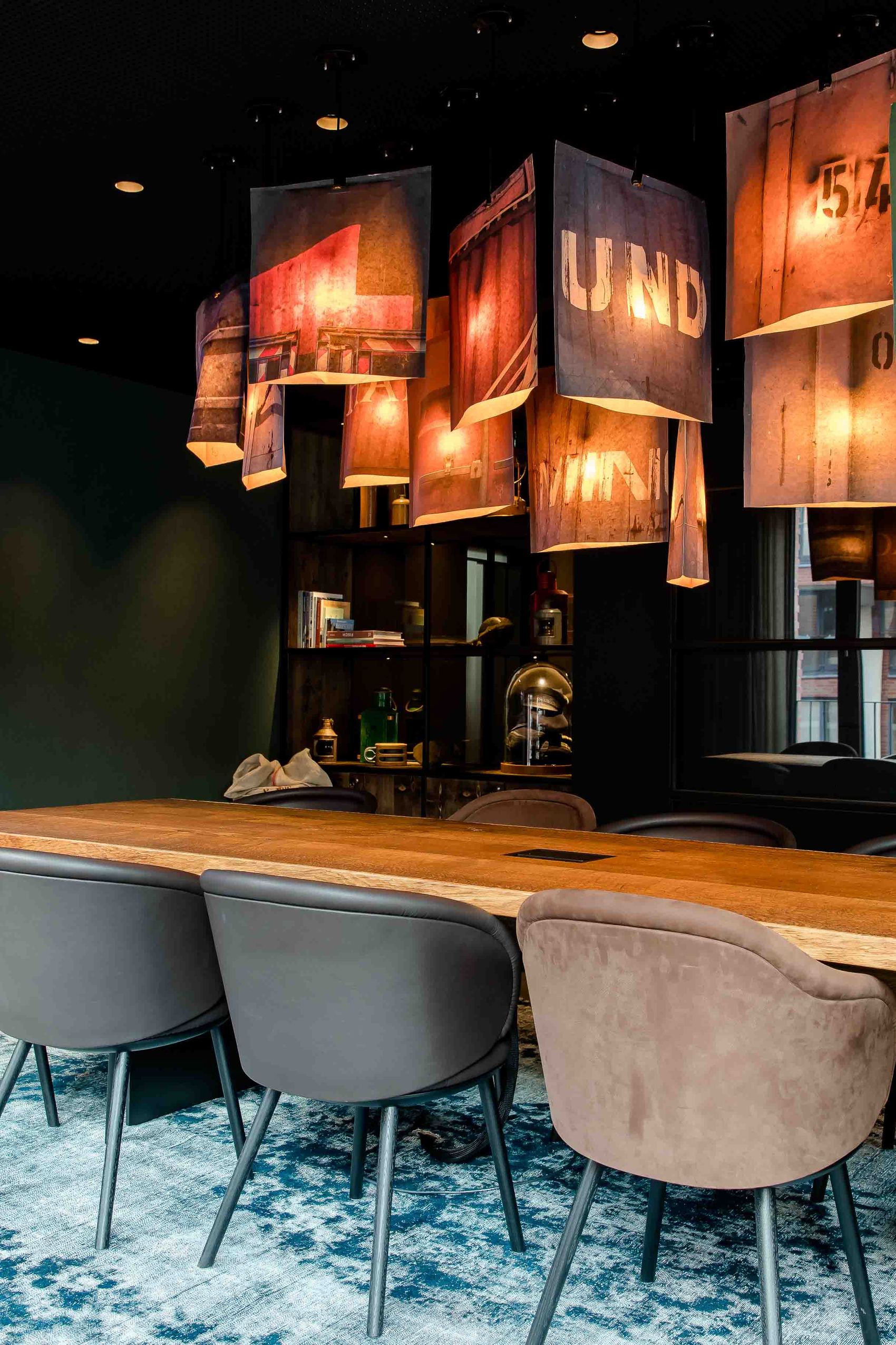 Motel One Besprechungsraum mit langem Holztisch, acht gepolsterten Stühlen und kreativen, beleuchteten Textillampen an der Decke. Ein großer Bildschirm zeigt ein Unterwasserbild.