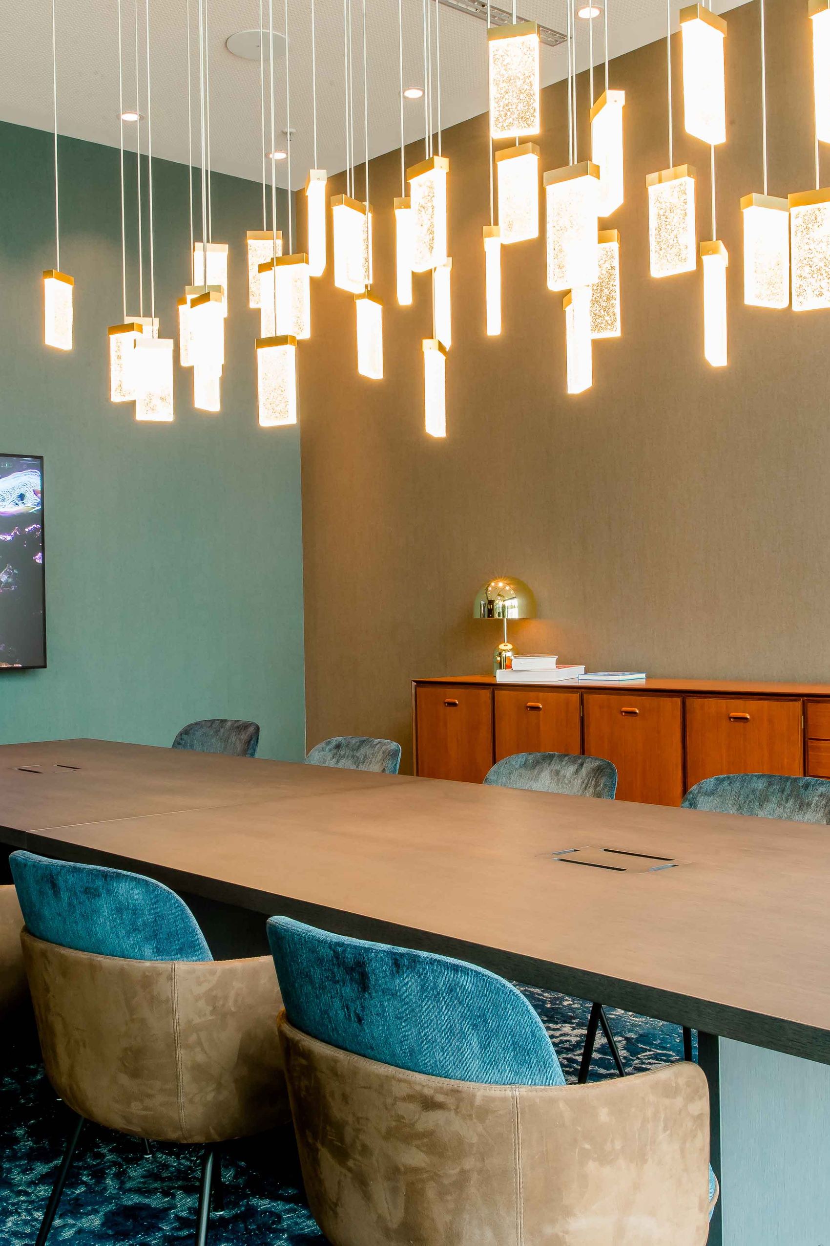 Motel One Konferenzraum mit langem Holztisch, moderner Beleuchtung und komfortablen Sesseln. Wandbild mit Unterwasser-Motiv und Sideboard mit dekorativer Lampe schaffen.