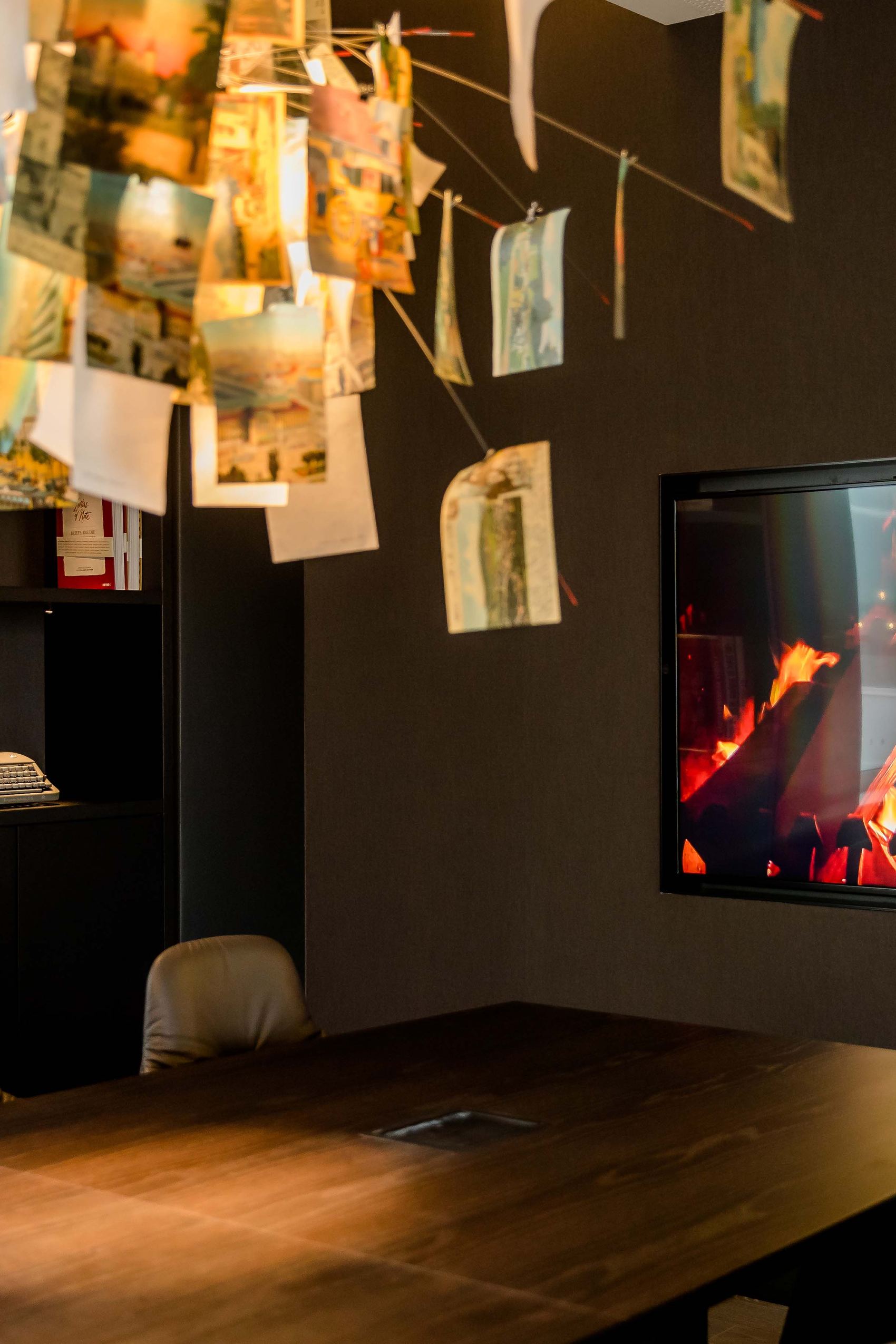 Motel One Besprechungsraum mit großem Holztisch, bequemen Stühlen und einem modernen Kaminfeuer an der Wand. Über dem Tisch hängt eine kreative, kunstvolle Deckenlampe aus vielen Papierblättern.