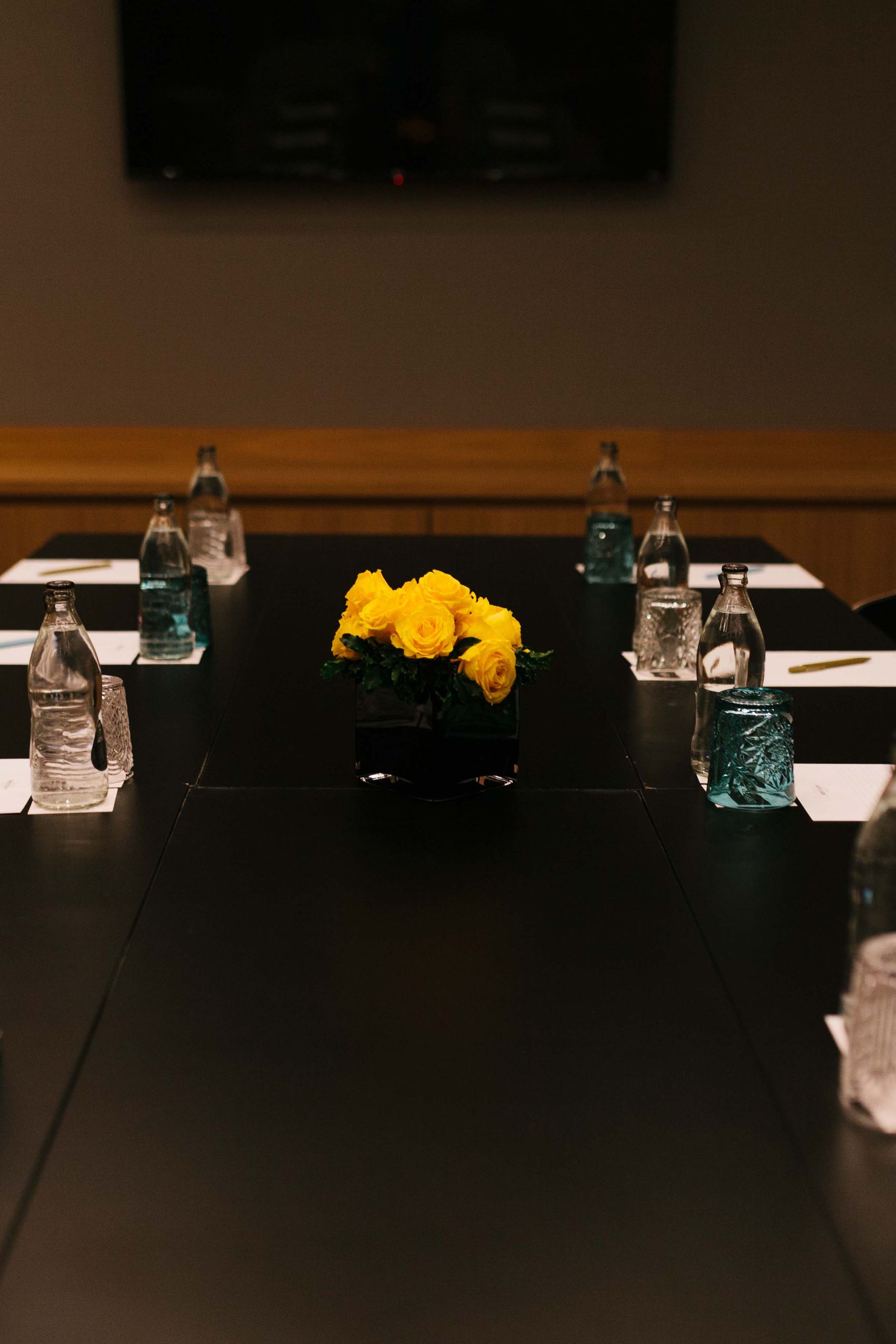 Motel One Konferenzraum mit langem Tisch, Sitzplätzen, Wasserflaschen, Schreibblöcken und Stiften, dekoriert mit gelben Blumen als Mittelpunkt, gedämpfte Beleuchtung für professionelle Atmosphäre.