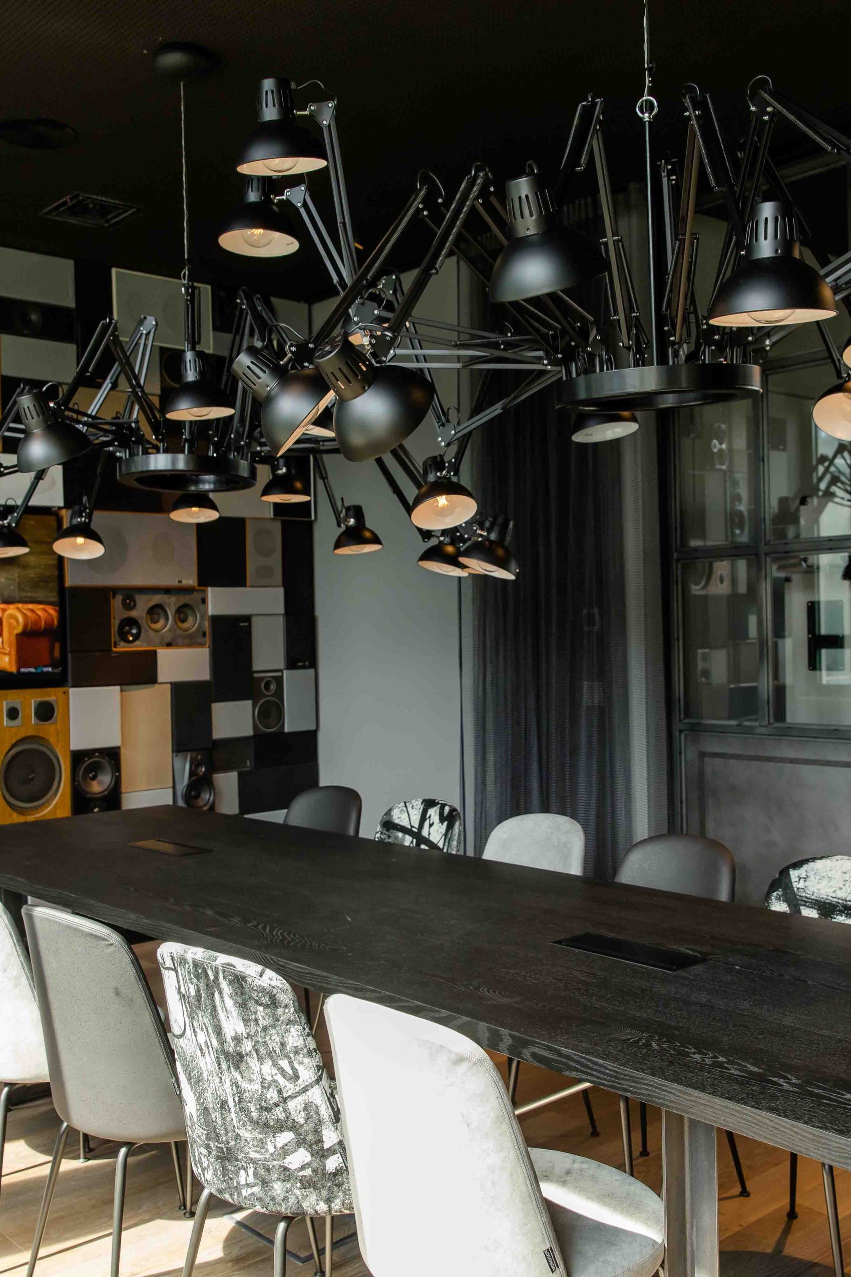 Motel One stilvoller Besprechungsraum mit großem dunklem Holztisch, acht verschieden gemusterte Stühle und kreativer Deckenbeleuchtung. Die Wand ist mit Lautsprechern und dekorativen Elementen gestaltet.