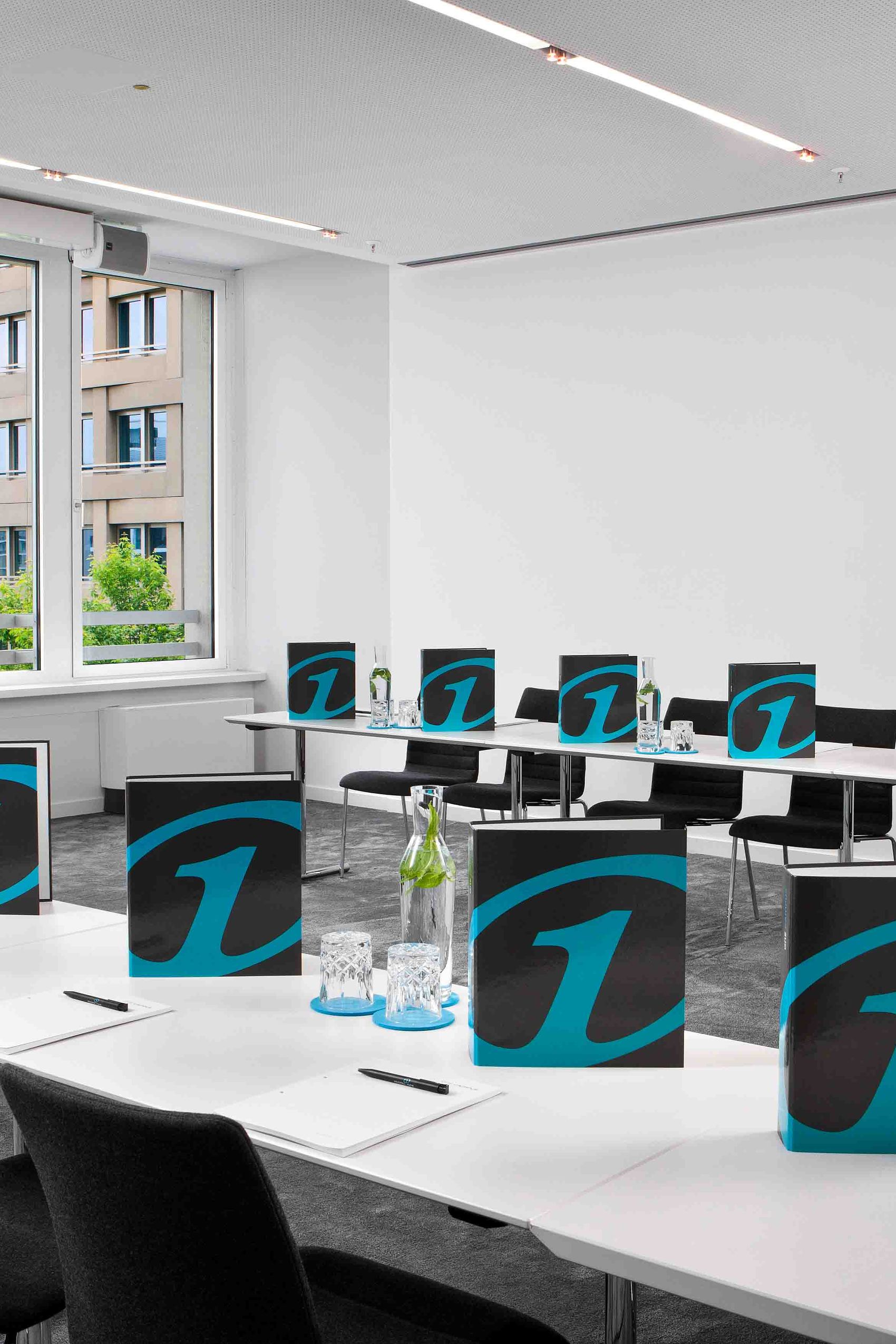 Motel One Konferenzraum mit großen Fenstern, Mobiliar und vorbereitetem Arbeitsplatz für mehrere Teilnehmer, ausgestattet mit Informationsmappen, Schreibblöcken und Wasserflaschen.