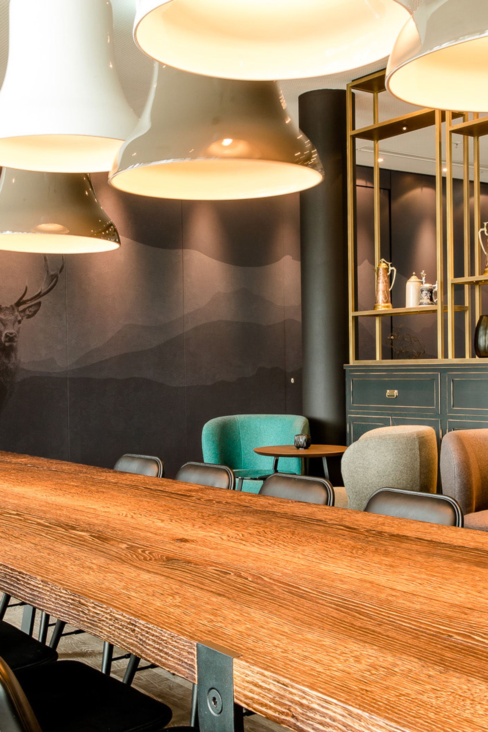 Motel One Konferenzraum mit langem Holztisch, bequemen Stühlen, stilvollen Hängelampen und einer Wandgestaltung mit Hirschmotiv.