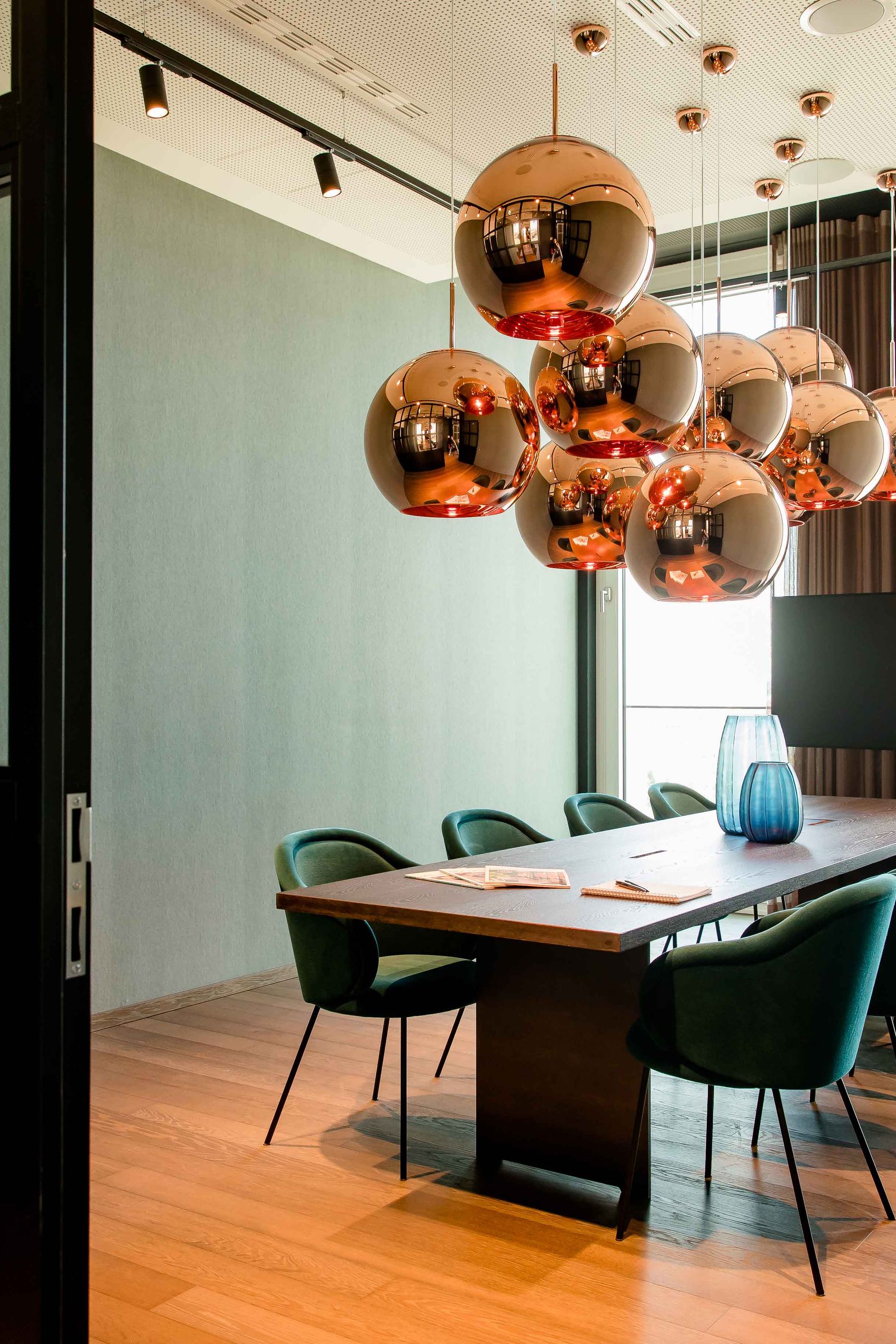 Motel One Konferenzraum mit langem Holztisch, acht grünen Stühlen und kupferfarbenen Kugelpendelleuchten, hell durch großflächige Fenster mit Tageslicht erleuchtet.