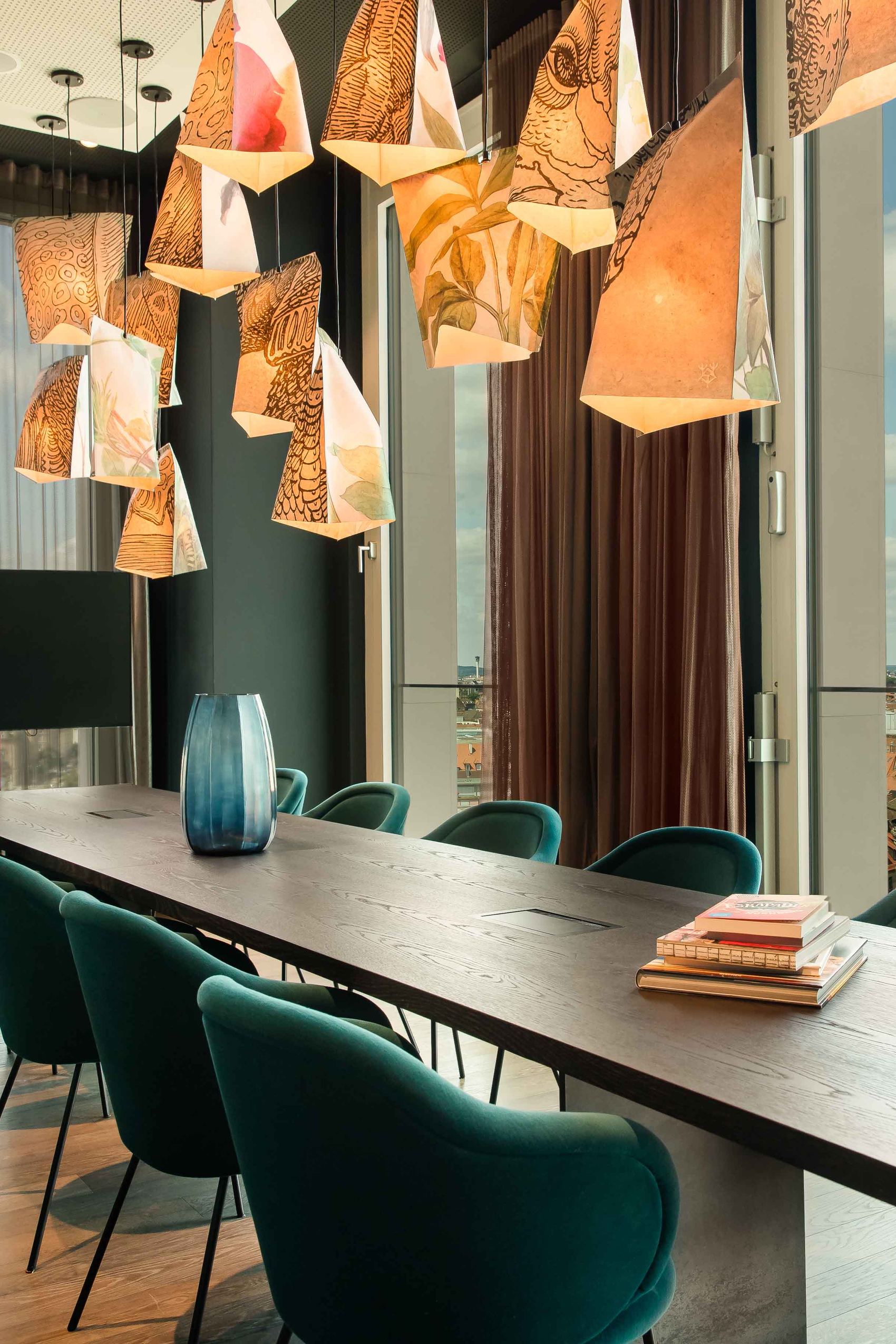 Motel One Besprechungsraum mit langem Holztisch, gemütlichen grünen Stühlen und dekorativen, warm beleuchteten Hängelampen. Große Fenster bieten Tageslicht und Ausblick auf die Stadt. Raum wirkt einladend und eignet sich für Geschäftstreffen oder kreative Gespräche.