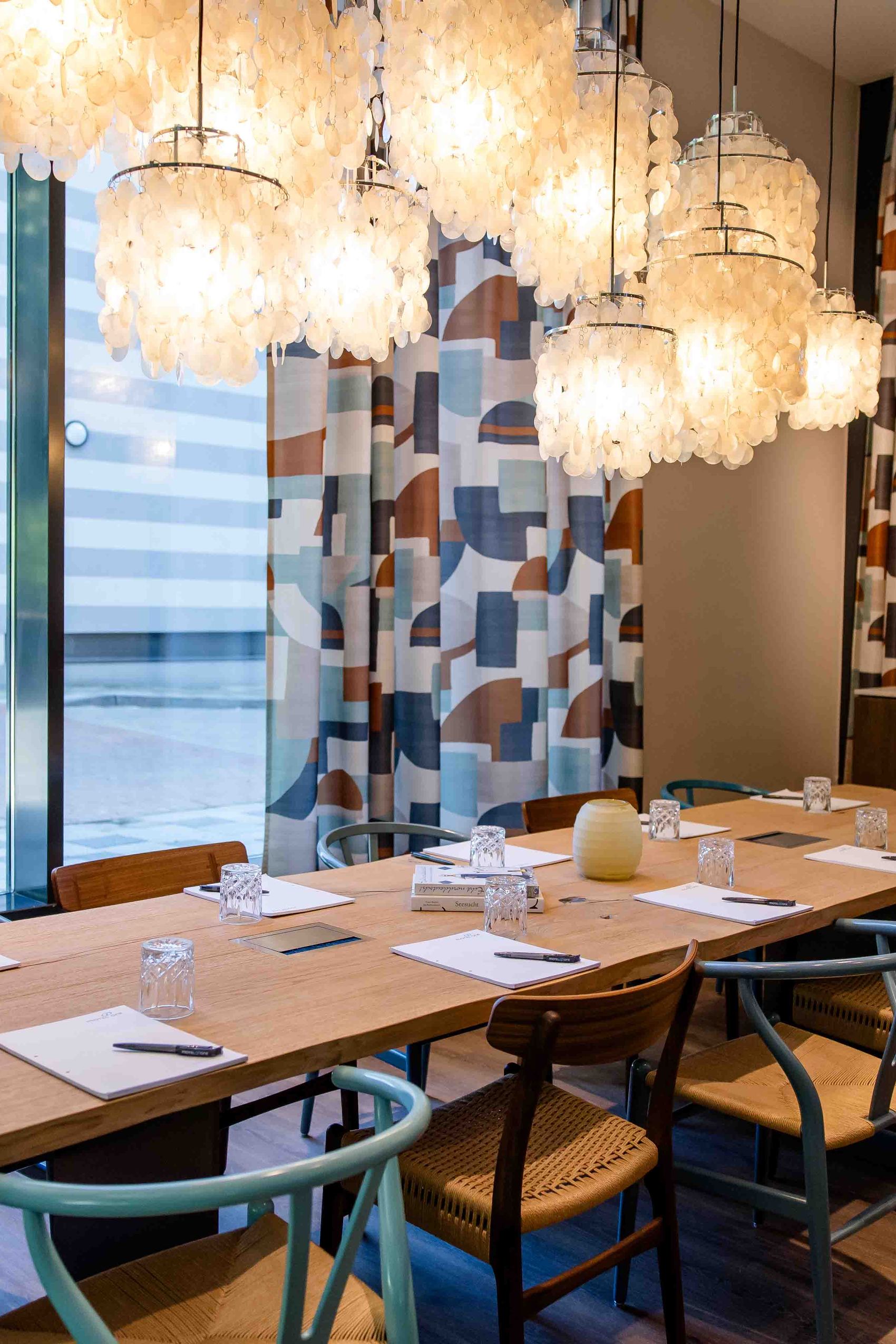 Motel One Konferenzraum mit großem Holztisch, bequemen Stühlen und stilvollen, leuchtenden Lampen. Große Fenster bieten Tageslicht und Blick auf die Stadt. Ideal für Besprechungen oder Gruppenarbeiten.