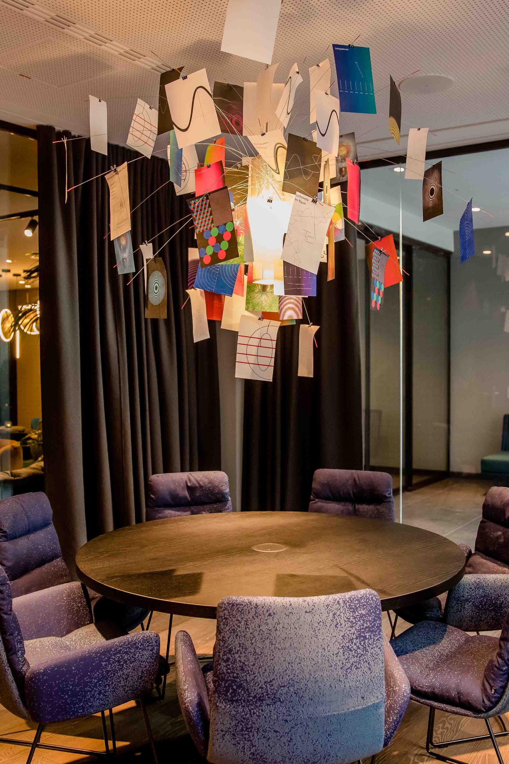 Motel One Besprechungszimmer mit rundem Holztisch, sechs gepolsterten Stühlen und kreativ gestalteter Deckenbeleuchtung aus hängenden Karten. Große Fenster und dunkle Vorhänge bieten Flexibilität bei Licht und Privatsphäre.