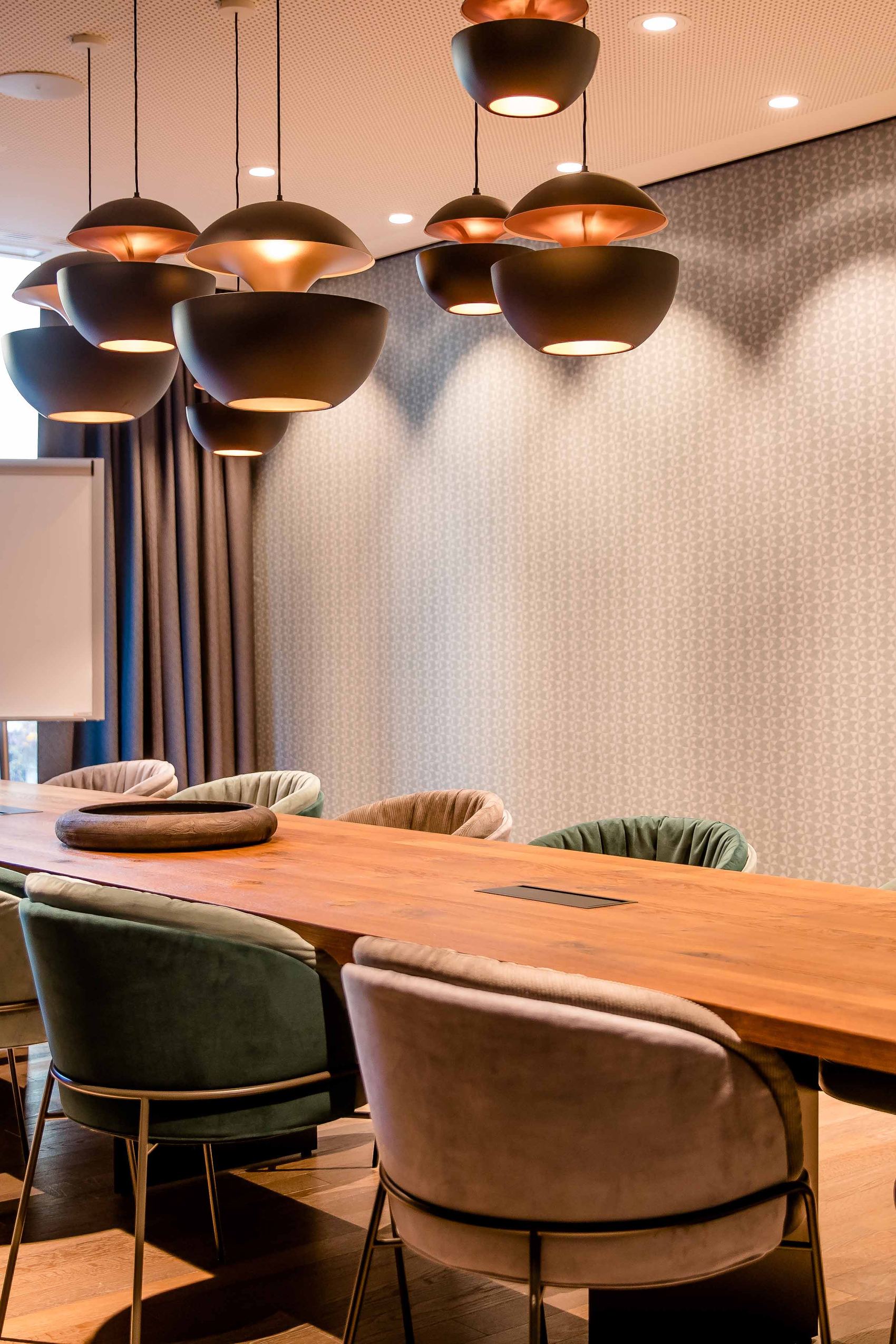 Motel One Konferenzraum mit großem Holztisch, komfortablen Stühlen und stilvollen Hängeleuchten.