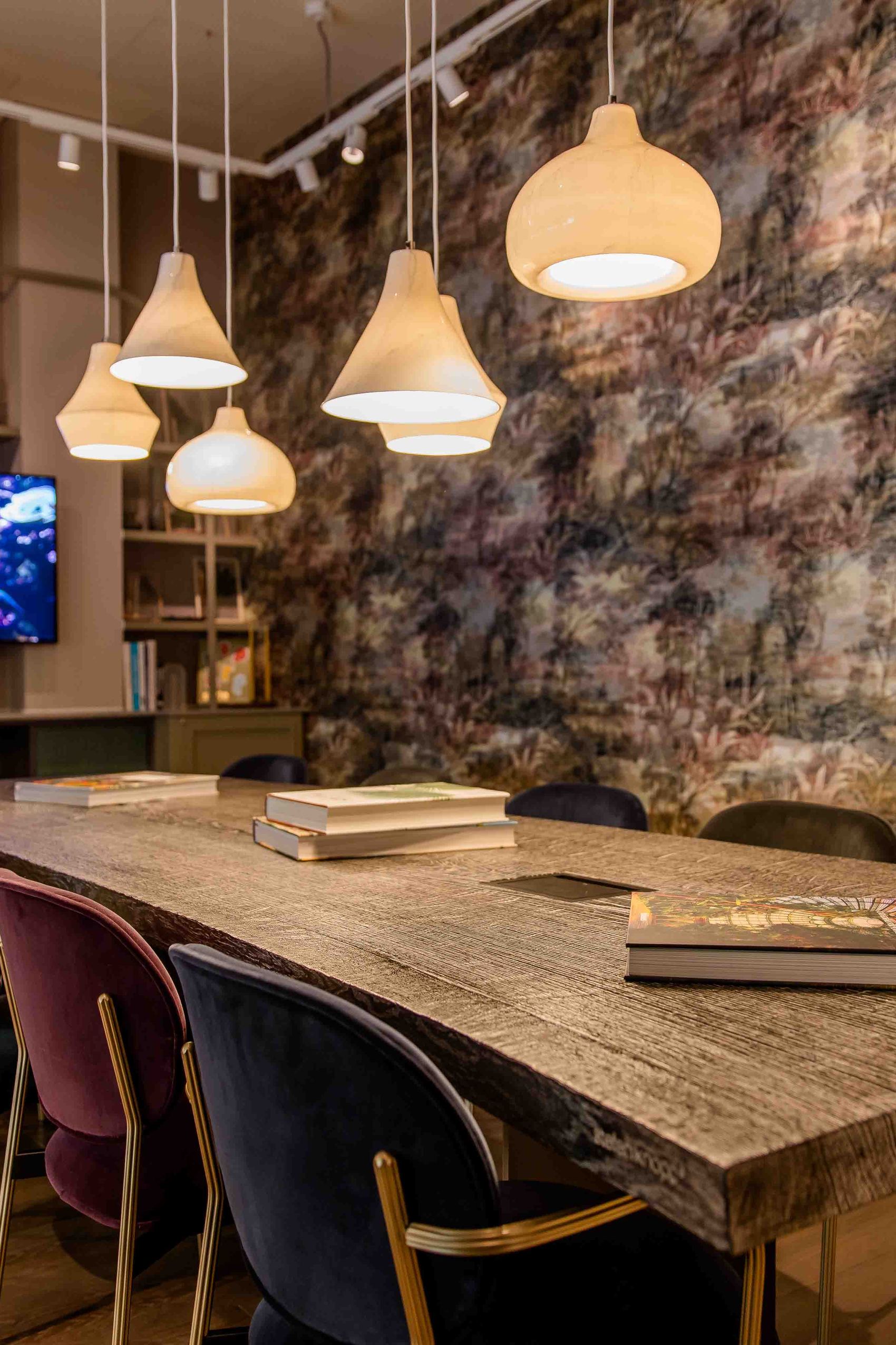 Motel One Besprechungsraum mit rustikalem Holztisch, bequemen Stühlen, warmem Licht durch mehrere Hängeleuchten und einer künstlerisch gestalteten Wand. Ein Bildschirm an der Wand zeigt eine Meeresaufnahme.