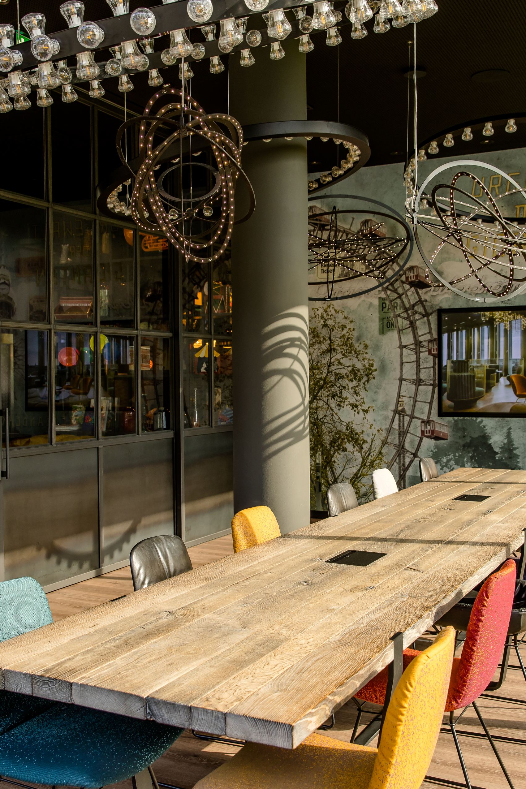 Motel One Besprechungsraum mit langem Holztisch, bunten Stühlen und großen Fenstern, die viel Tageslicht hereinlassen. An der Wand Kunstobjekte und ein Bildschirm für Präsentationen.