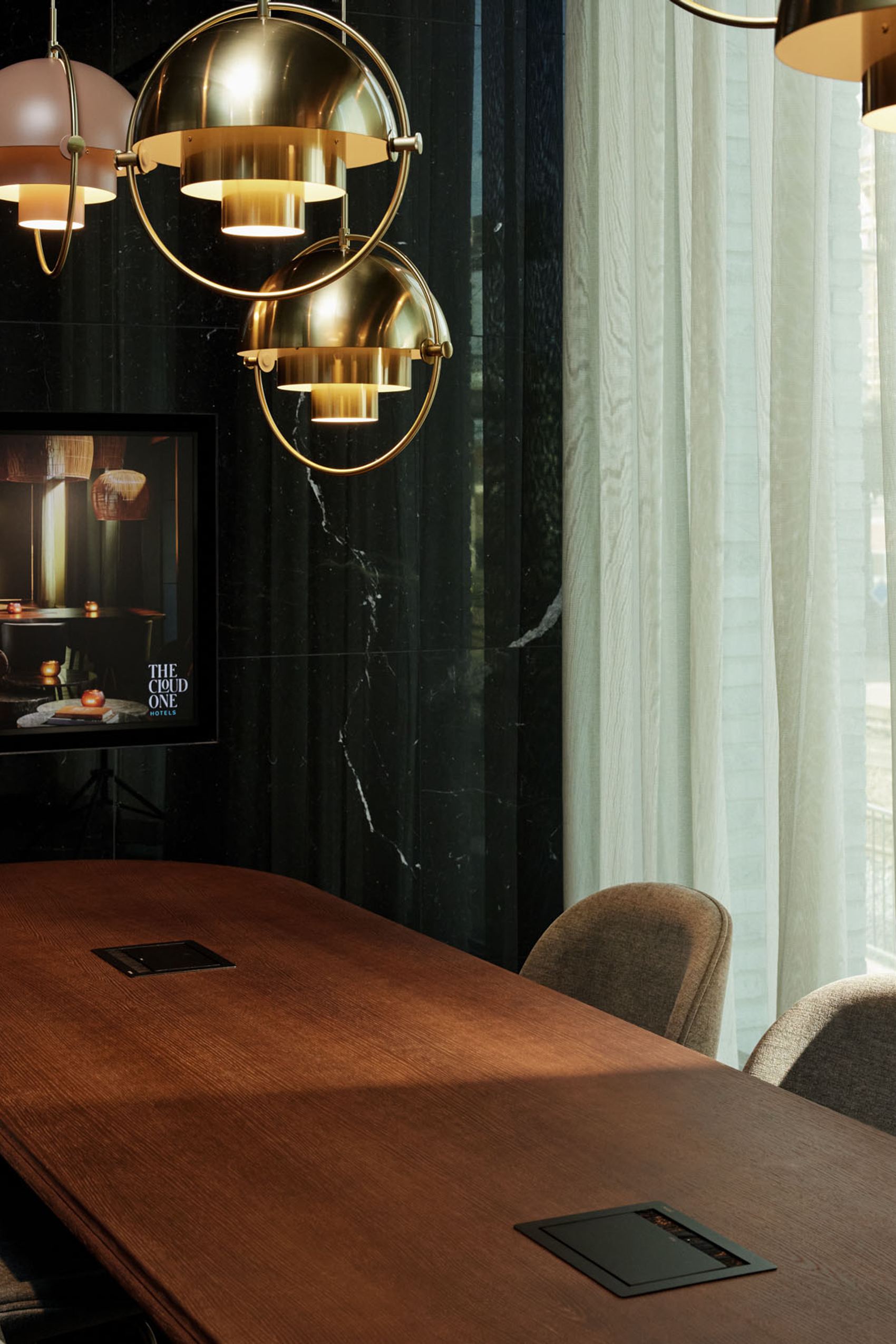 Motel One Konferenzraum mit langem Holztisch, bequemen Stühlen und großen Fenstern, die Tageslicht hereinlassen. Stilvolle goldene Hängeleuchten pendeln von der Decke.