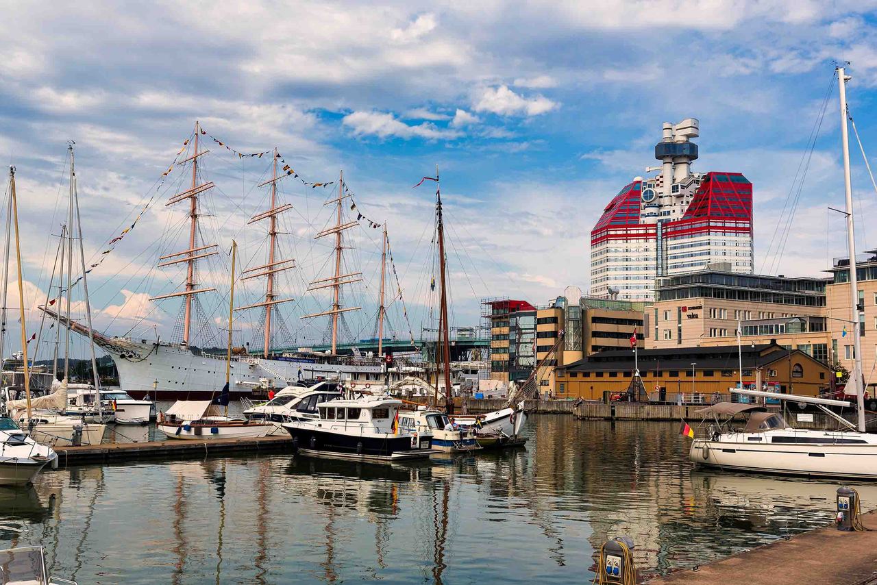 Hafen mit Segel- und Motorbooten im Wasser, daneben Gebäude, darunter ein auffälliges rot-weißes Hochhaus.