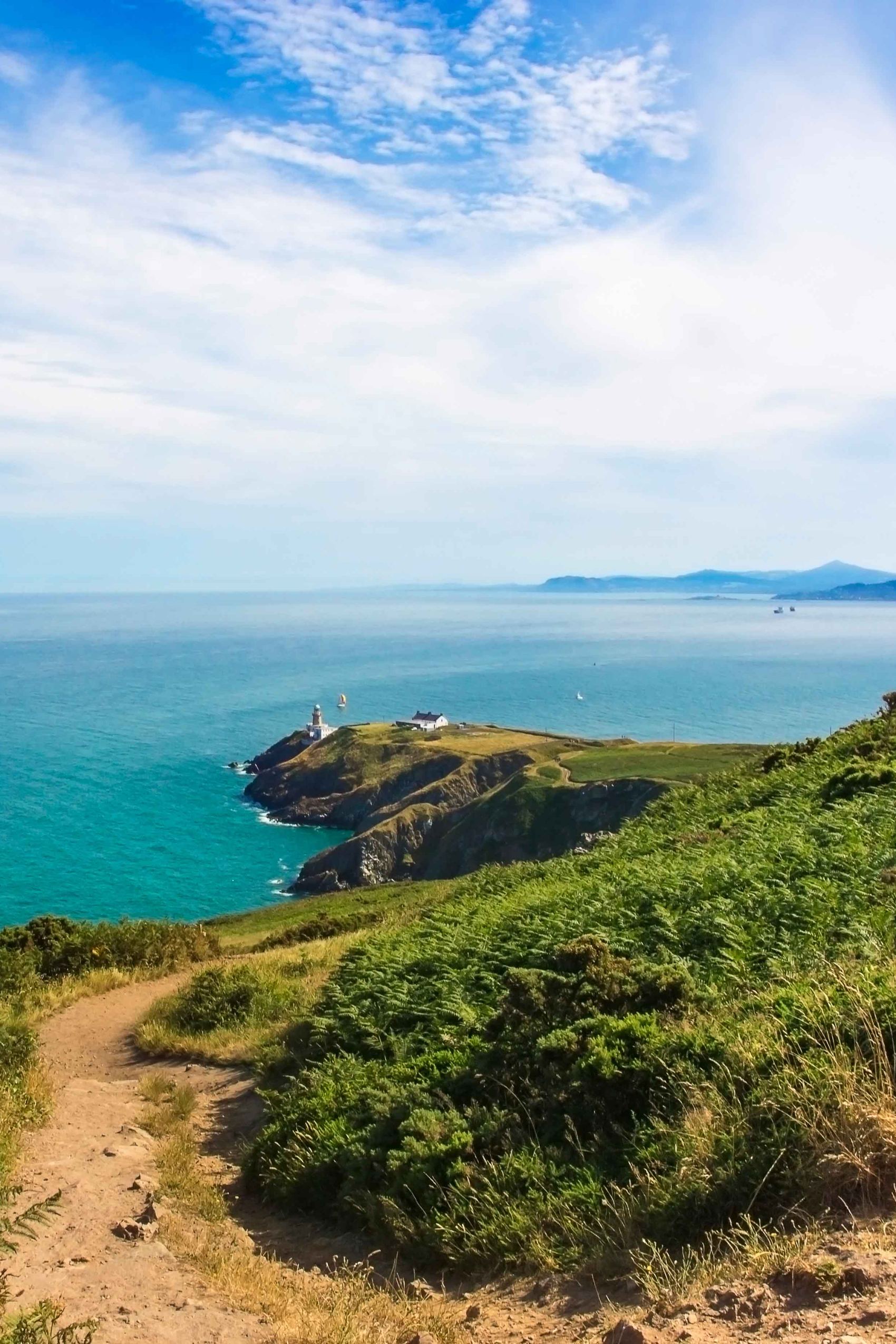 Blick auf einen Küstenweg entlang grüner Hügel mit Blick auf ein Kap und einen Leuchtturm, umgeben vom blauen Meer unter einem teils bewölkten Himmel.