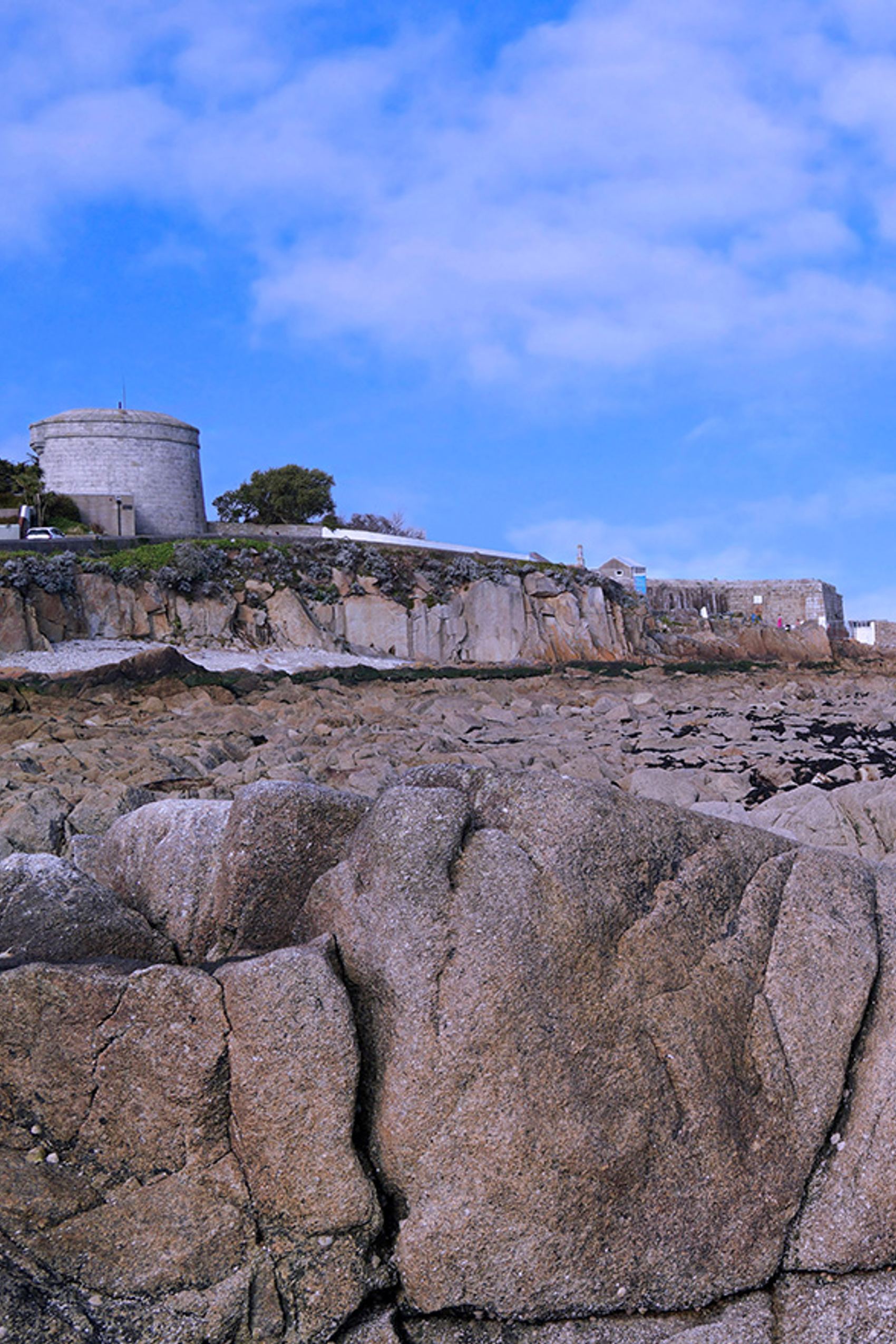 Felsige Küstenlandschaft unter blauem Himmel mit einem historischen, rundlichen Steinturm auf einer Klippe, der eine ruhige, maritime Atmosphäre vermittelt.