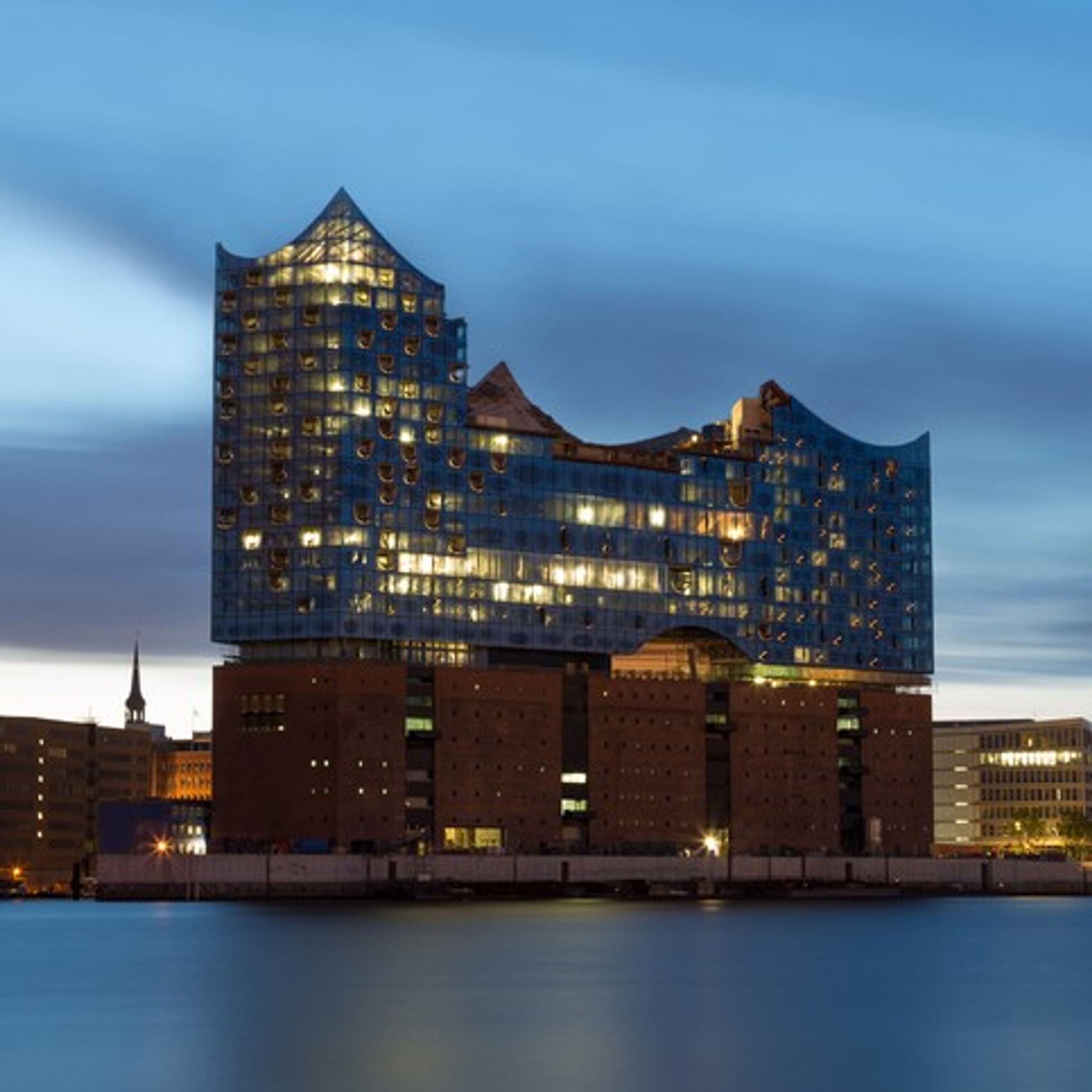 Moderne Elbphilharmonie in Hamburg bei Dämmerung, gläserne Wellenstruktur auf Ziegelsteinsockel reflektiert Lichter, sanfter Himmel und ruhiges Wasser im Vordergrund.
