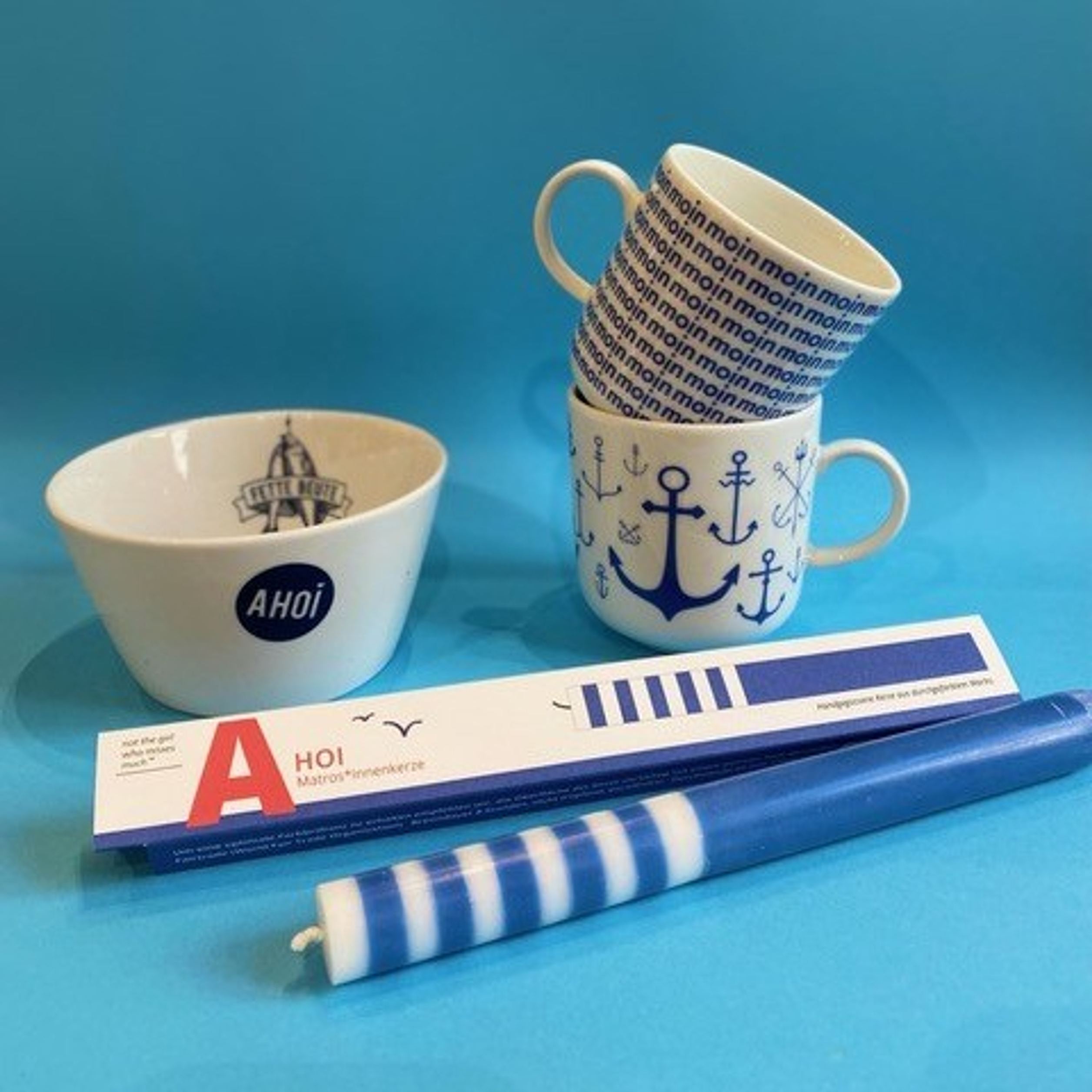 Weiße Tassen mit maritimen Motiven wie Ankern und dem Schriftzug „moin“ sowie eine Schale mit dem Wort „AHOI“. Dazu eine blaue Kerze mit weißen Streifen, alles vor blauem Hintergrund arrangiert, vermittelt nordische Gemütlichkeit.