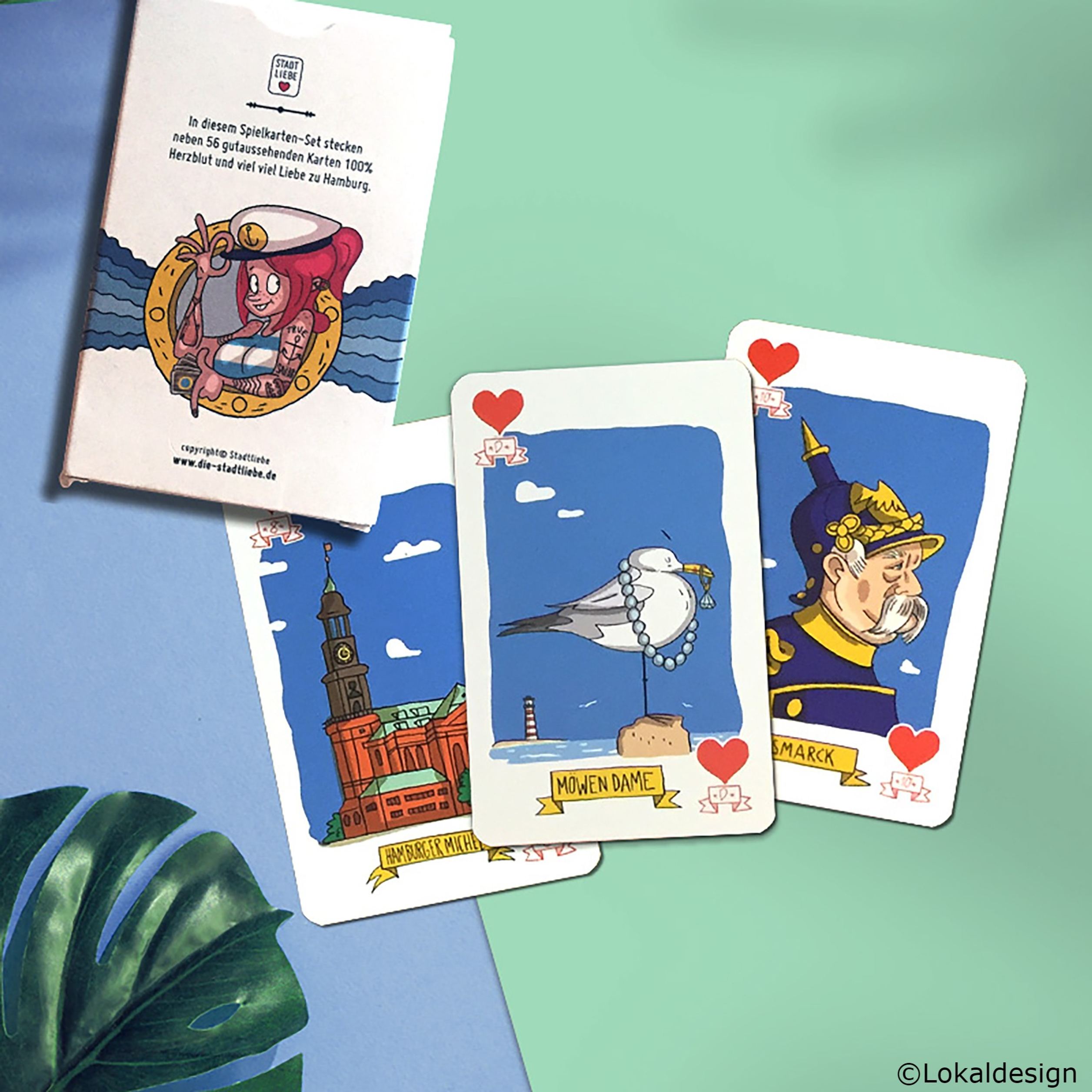 Spielkarten mit freundlichen Illustrationen bekannter Hamburger Wahrzeichen und Figuren, darunter Mönkedamm-Seemöwe, Rathaus und Bismarck, auf farbigem Hintergrund. Verpackung erwähnt die Lokalbezüge und enthält 56 Karten mit Liebe zu Hamburg.