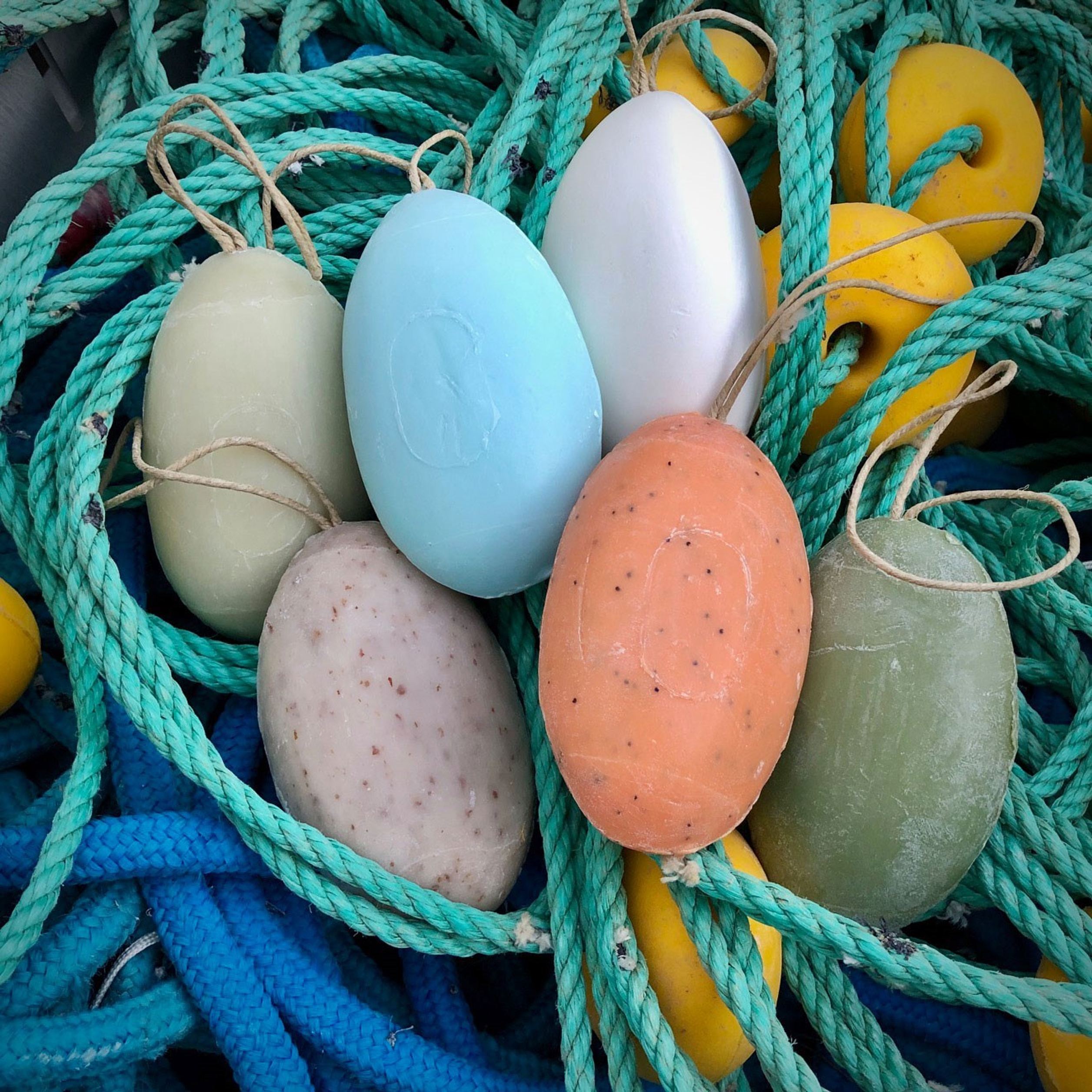 Sechs landestypische schwimmfähige Seifen in verschiedenen Farben, präsentiert auf einem blauen und grünen Tauwerksnetz mit gelben Schwimmern, symbolisieren maritime Tradition und Handwerkskunst.