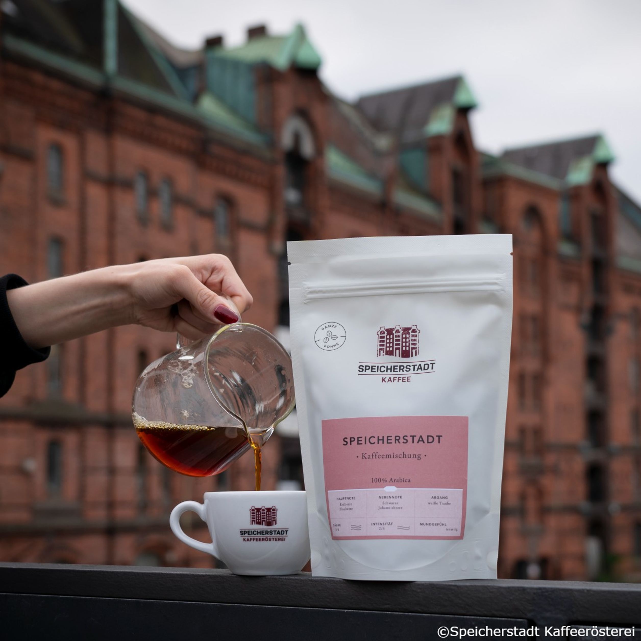 Hand gießt frisch gebrühten Kaffee aus Glaskanne in Tasse mit Logo „Speicherstadt Kaffeerösterei“. Daneben steht ein Beutel Kaffee „Speicherstadt Kaffee“ vor historischer Backsteinfassade.