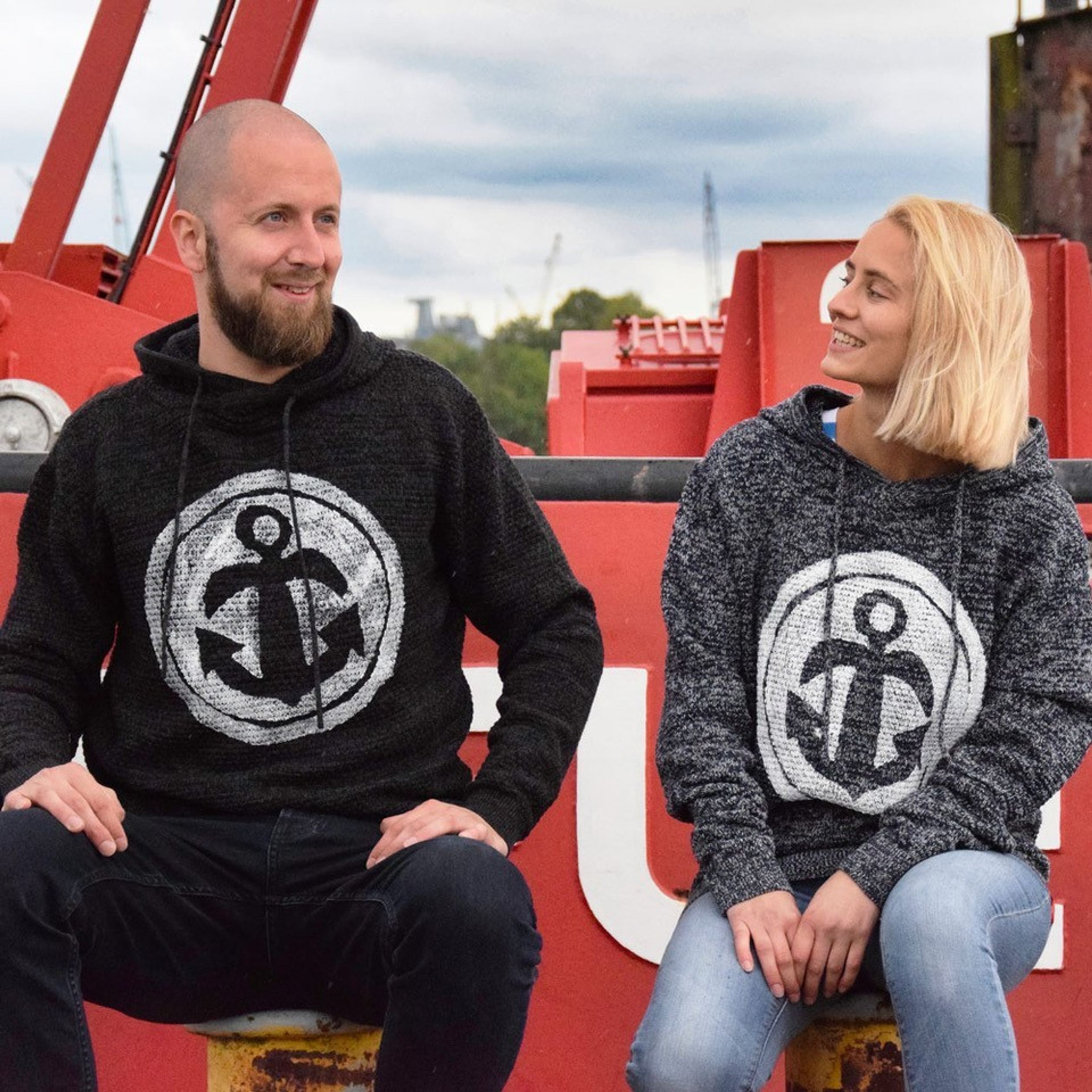 Zwei Personen sitzen entspannt nebeneinander, tragen Kapuzenpullover mit großem Anker-Symbol, lächeln sich an. Hintergrund deutet auf maritimes Umfeld hin.