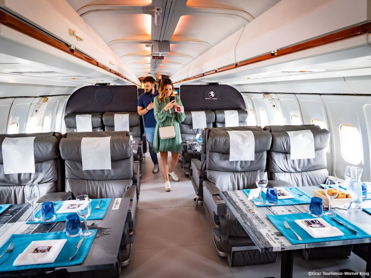 Innenansicht eines luxuriösen Flugzeuges mit großzügigen Ledersesseln und gedeckten Tischen, zwei Passagiere gehen den Gang entlang.
