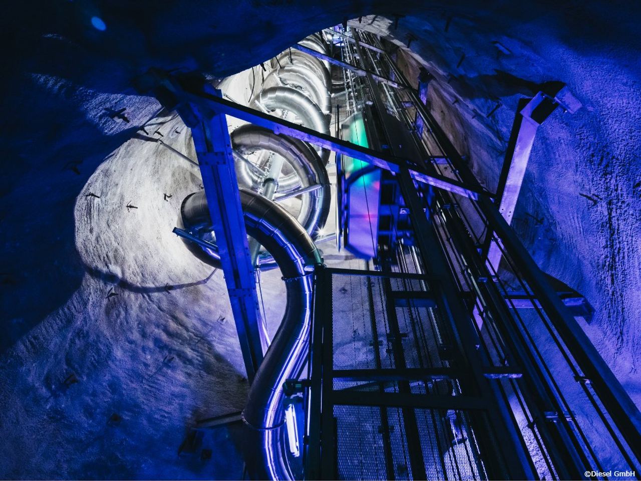 Blick in einen beleuchteten Tunnel mit metallischen Röhren und Strukturen, die durch Felswände verlaufen, in kühlem blauem Licht. Die Konstruktion wirkt technisch und industriell.
